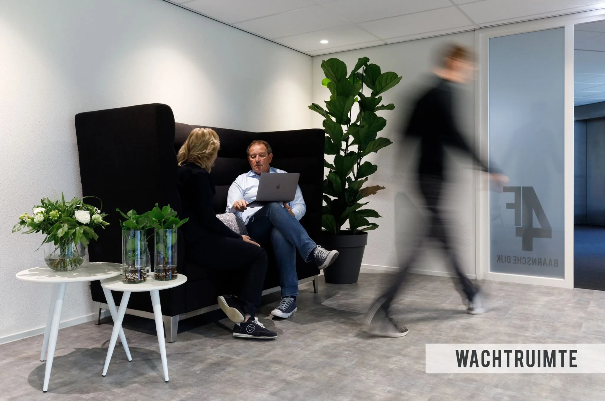 Twee mensen zitten in een moderne wachtkamer met planten en een laptop, bij een deur met het logo van 4E Baarnsche Dijk.
