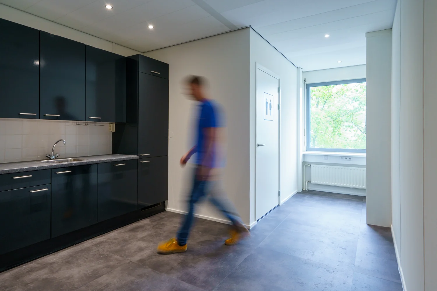 Man loopt door lichte gang langs een donkere kitchenette in een gebouw aan de Baarnsche Dijk.
