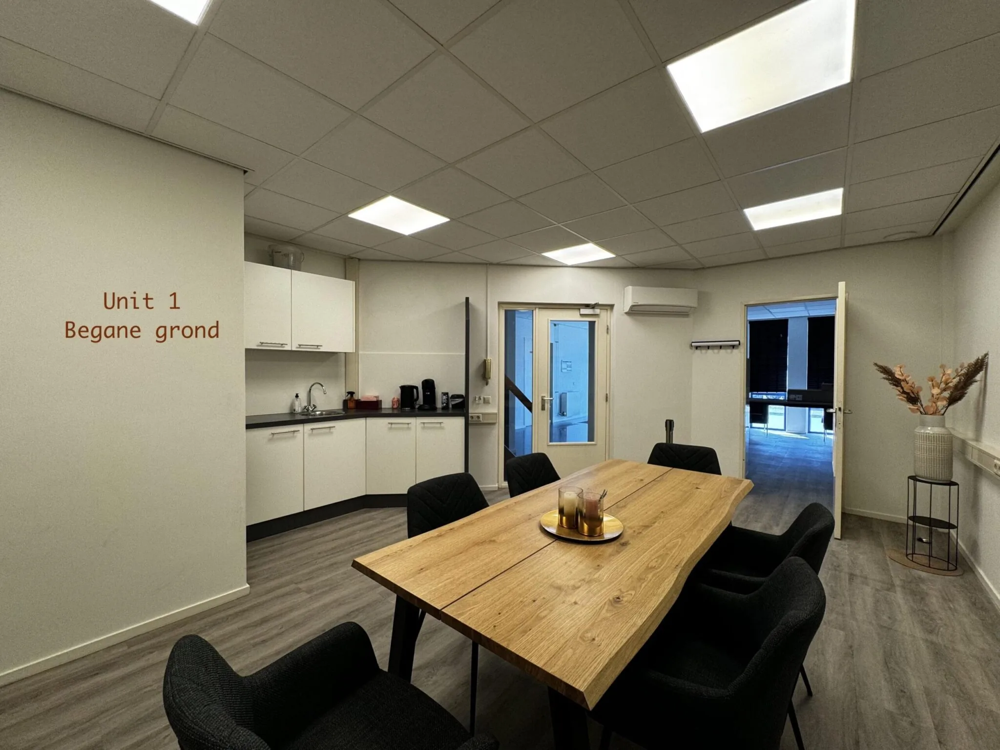 Vergaderruimte met houten tafel, zwarte stoelen, kitchenette en opschrift "Unit 1 Begane grond" op de muur.