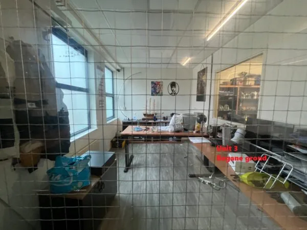 Kantoorruimte met bureaus, modelboot en decoratie, gezien door een roosterraam met rode tekst "Unit 3 Begane grond".