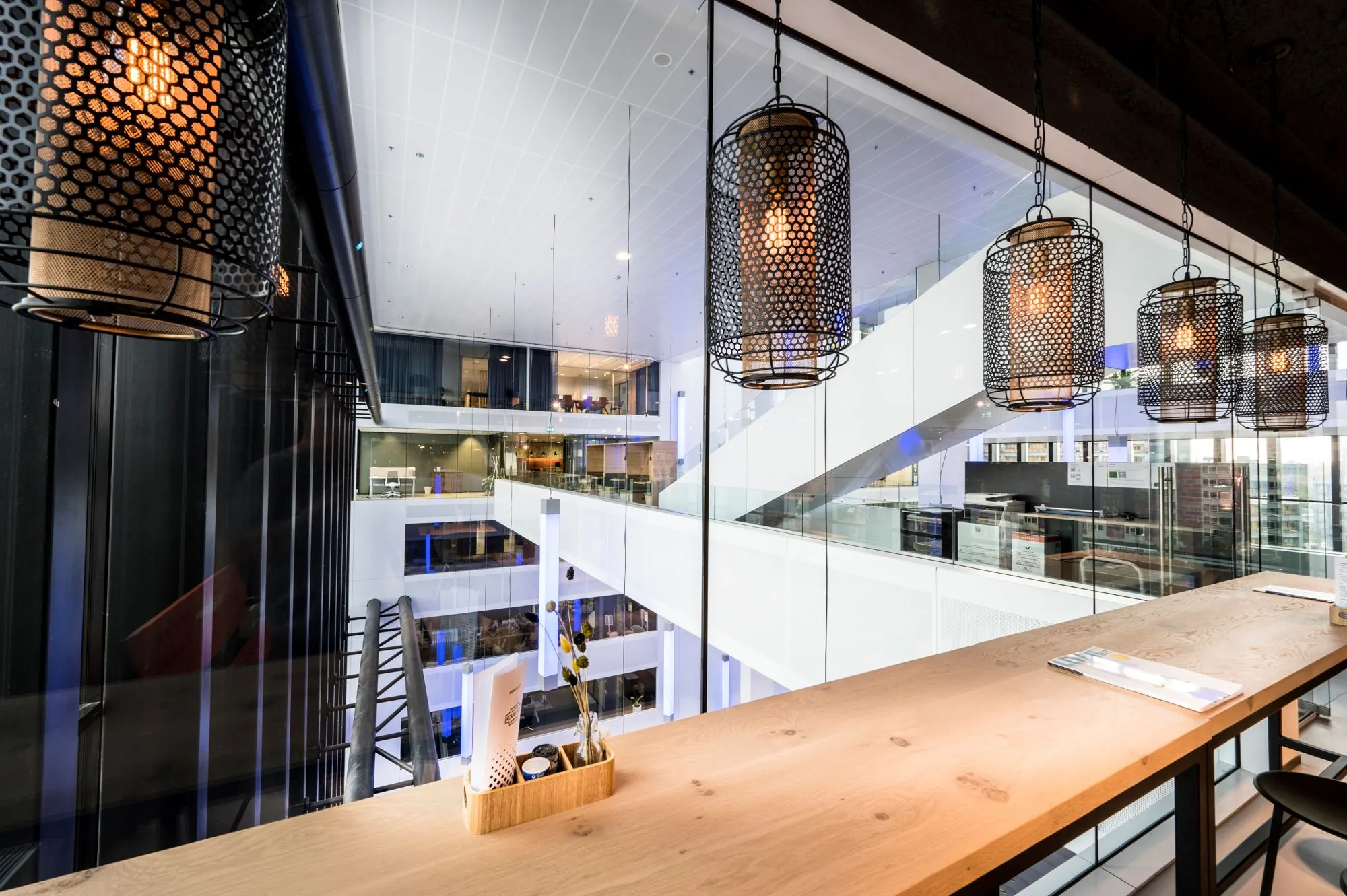 Moderne kantoorruimte met uitzicht op een open atrium en hangende industriële lampen boven een houten werktafel.