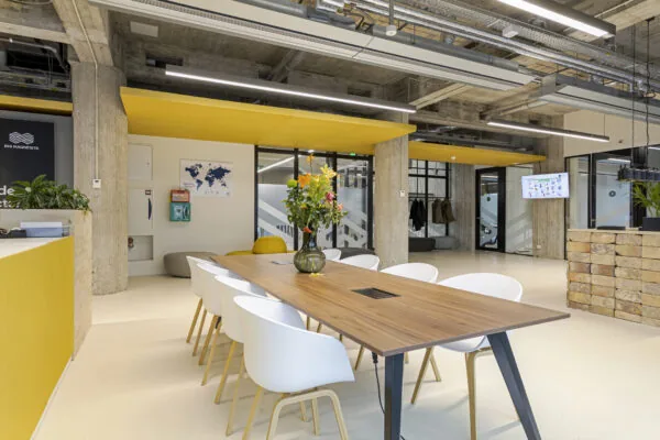 Moderne kantoorruimte met een lange tafel, witte stoelen, bloemenvaas en gele accenten.