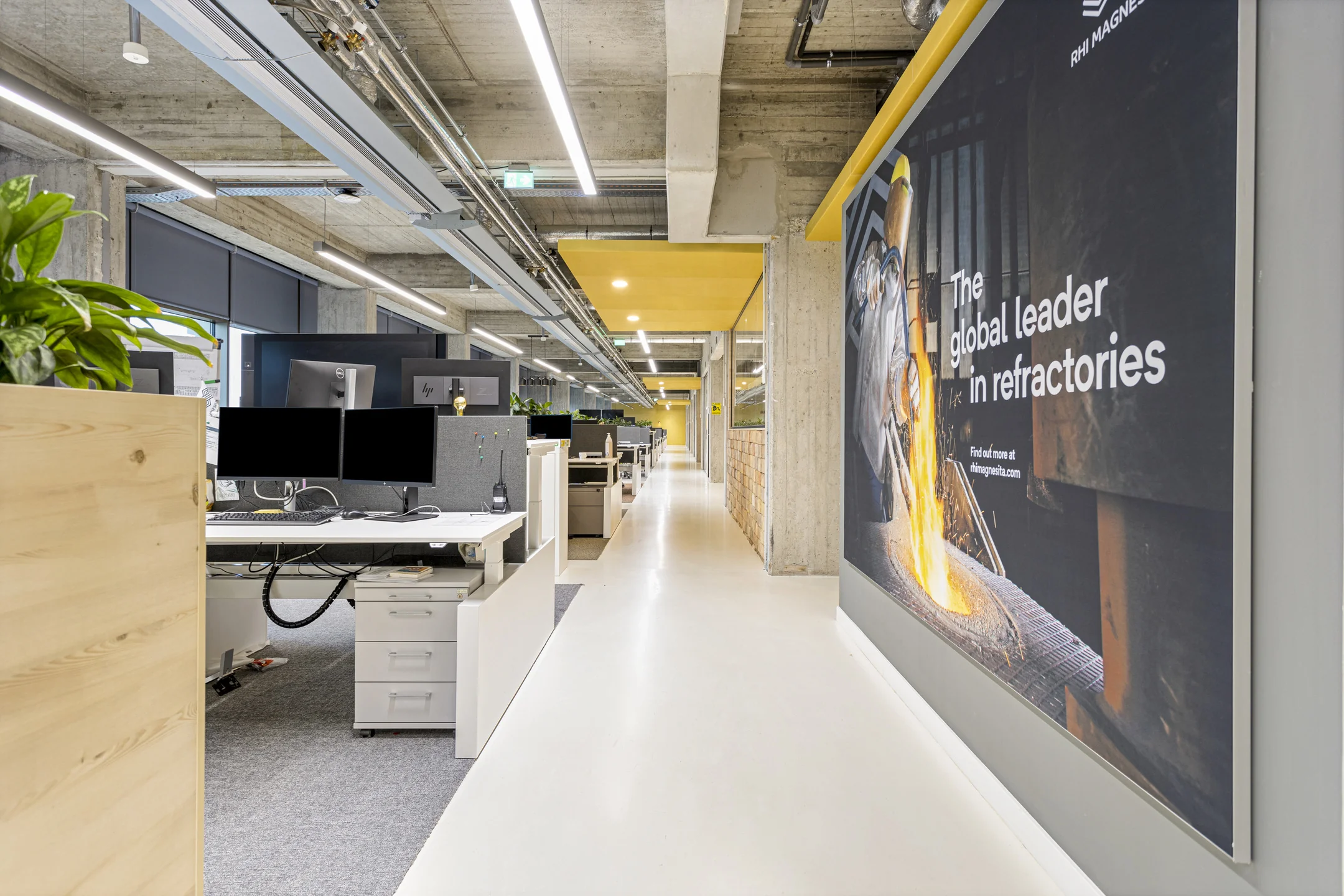 Modern open kantoorinterieur met werkplekken en een groot promotiebord van RHI Magnesita aan de muur.