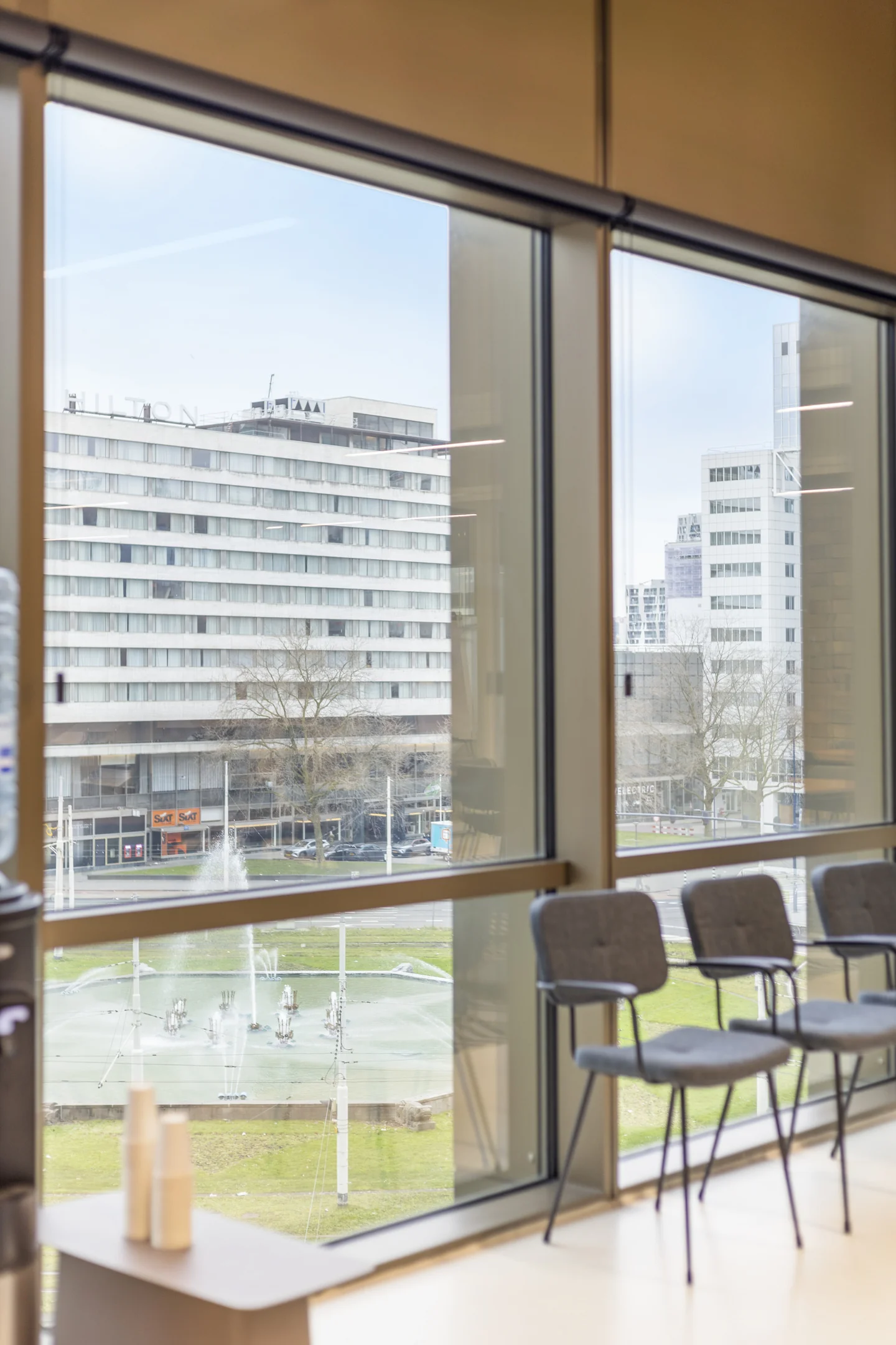 Uitzicht op het Hofplein met fonteinen vanuit een moderne kantoorruimte met stoelen en een waterkoeler bij het raam.