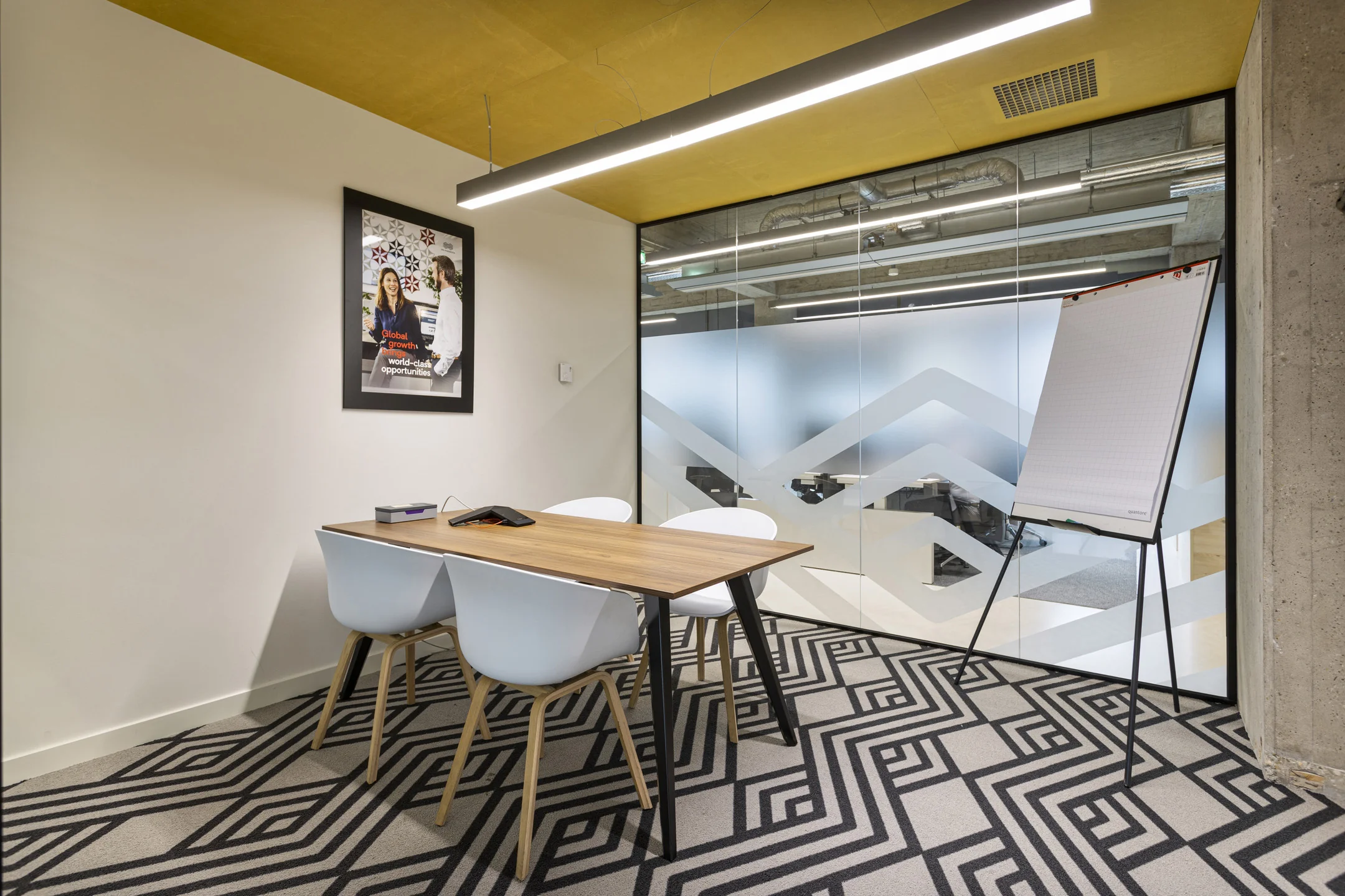 Een moderne vergaderruimte met houten tafel, witte stoelen, flip-over en een glazen wand met patroon.