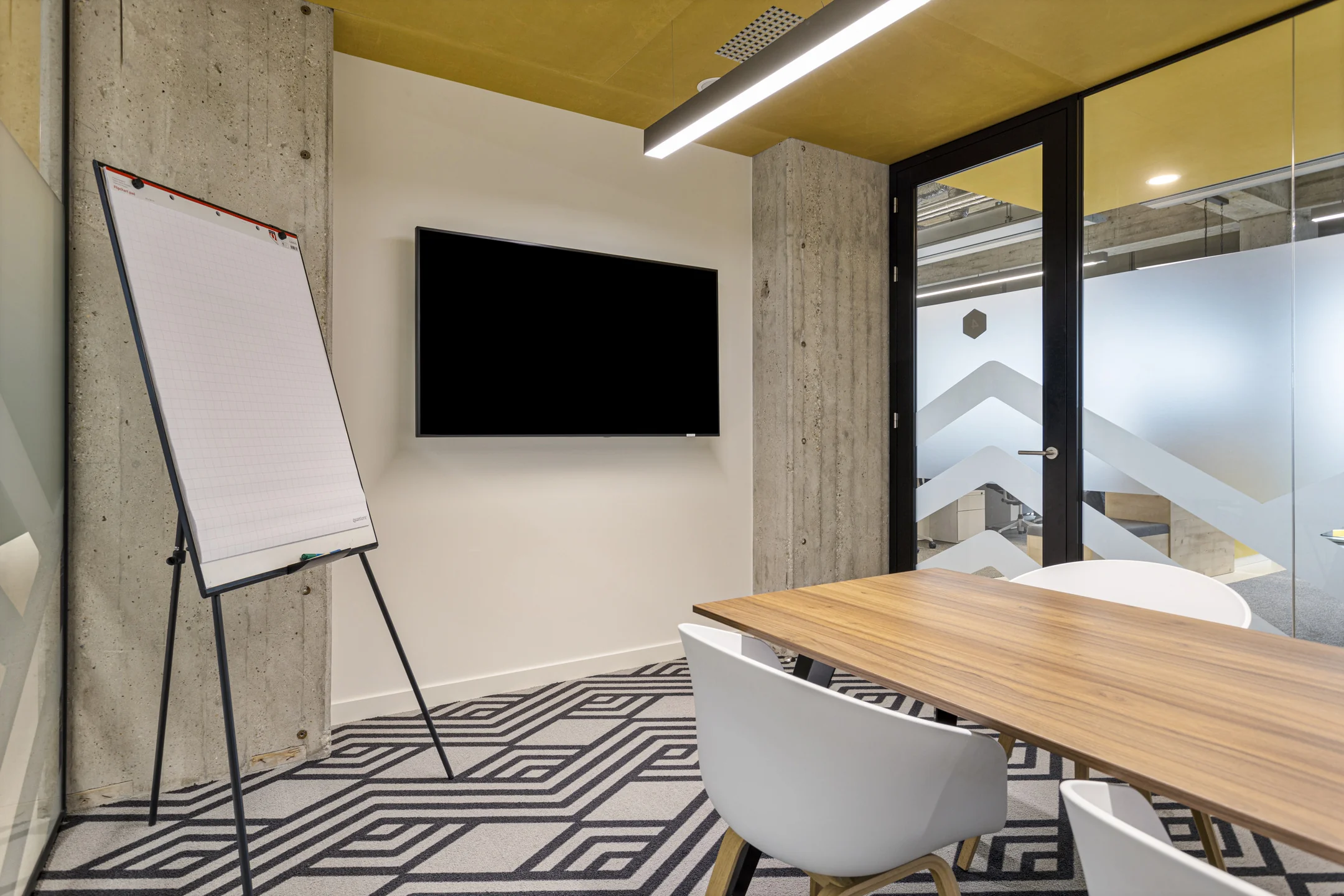Een moderne vergaderruimte met een flip-over, televisiescherm en houten tafel met witte stoelen.