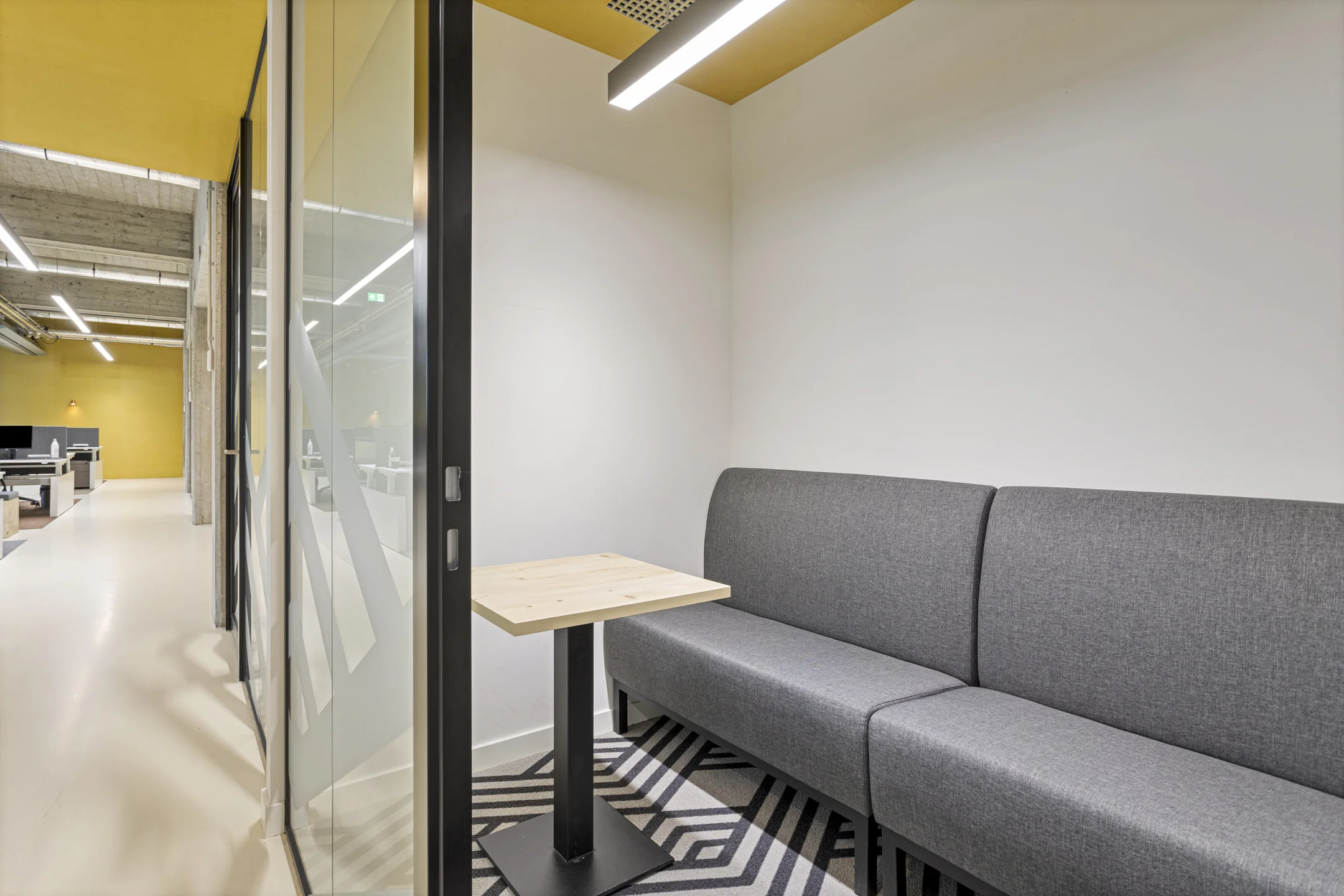 Moderne kantoorruimte met een grijze bank en houten tafel in een afgescheiden hoek, naast een open kantoorgedeelte.