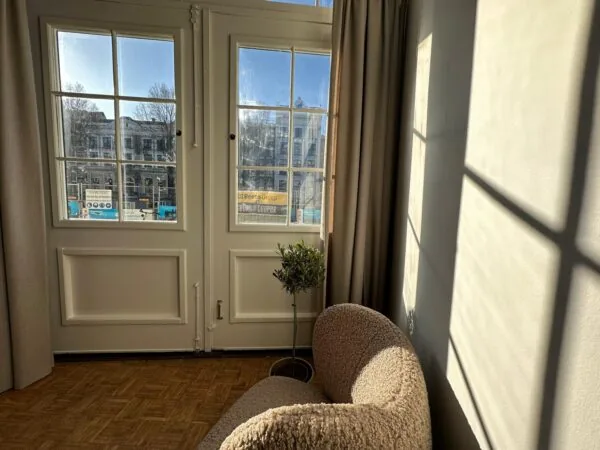 Zonnige kamer met uitzicht op de Herengracht, met een gebogen stoel en schaduw van raamkozijnen op de muur.