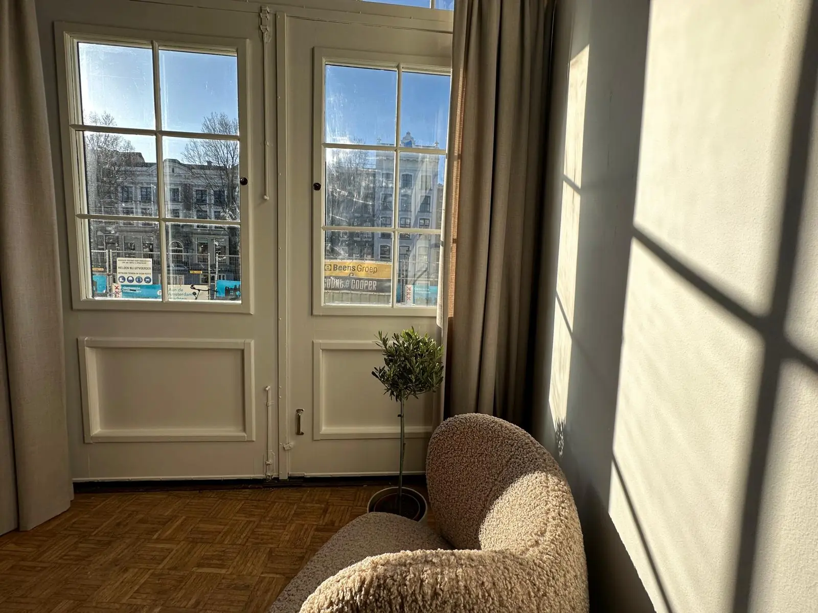 Zonnige kamer met uitzicht op de Herengracht, met een gebogen stoel en schaduw van raamkozijnen op de muur.