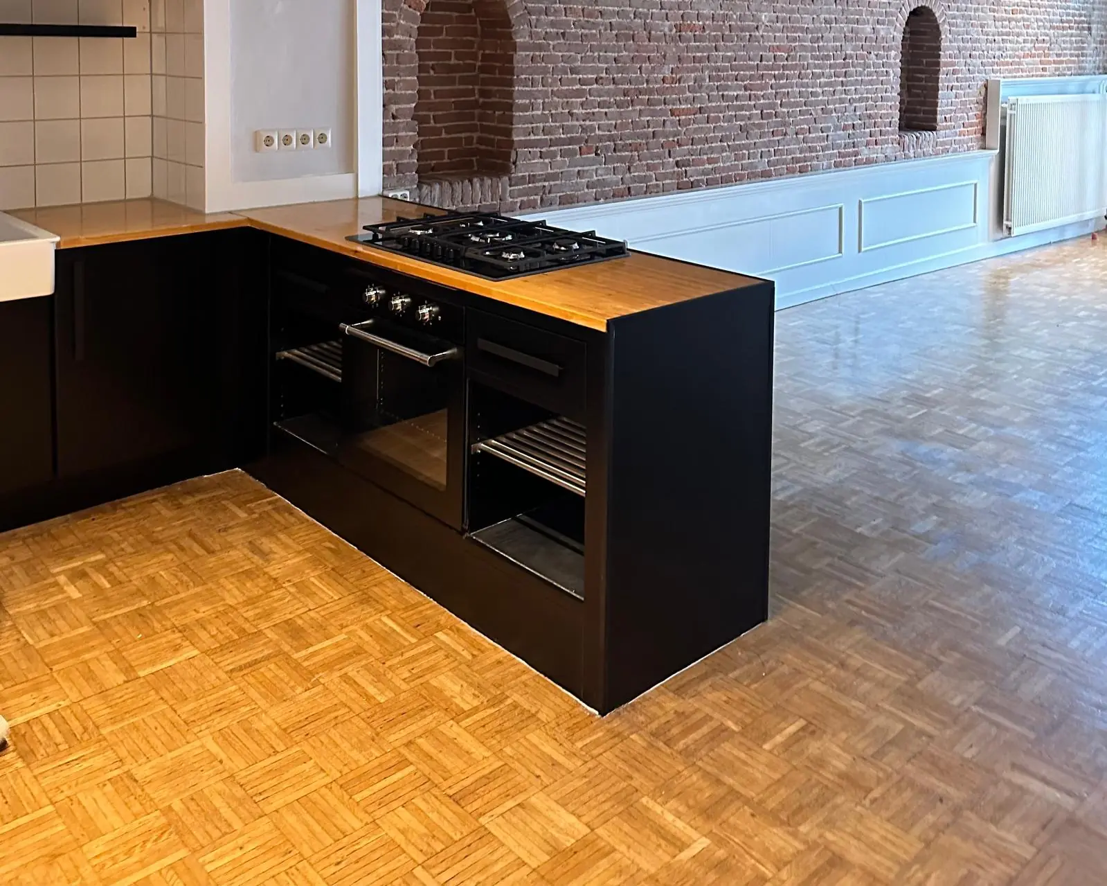 Moderne zwarte hoekkeuken met houten werkblad en inbouwoven in een appartement aan de Herengracht met karakteristieke bakstenen muur.
