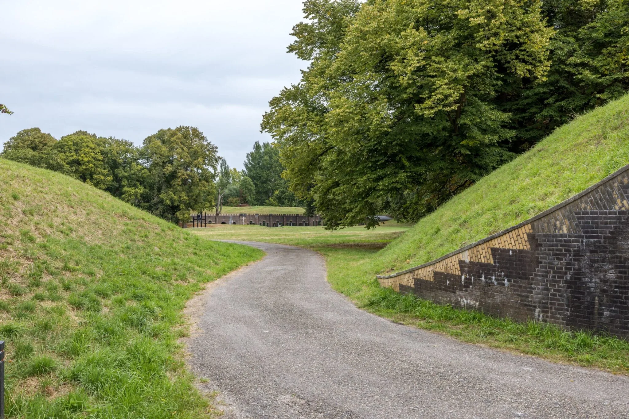 Geasfalteerd pad tussen met gras begroeide dijken bij de Vossegatsedijk met op de achtergrond een historisch fortgebouw.
