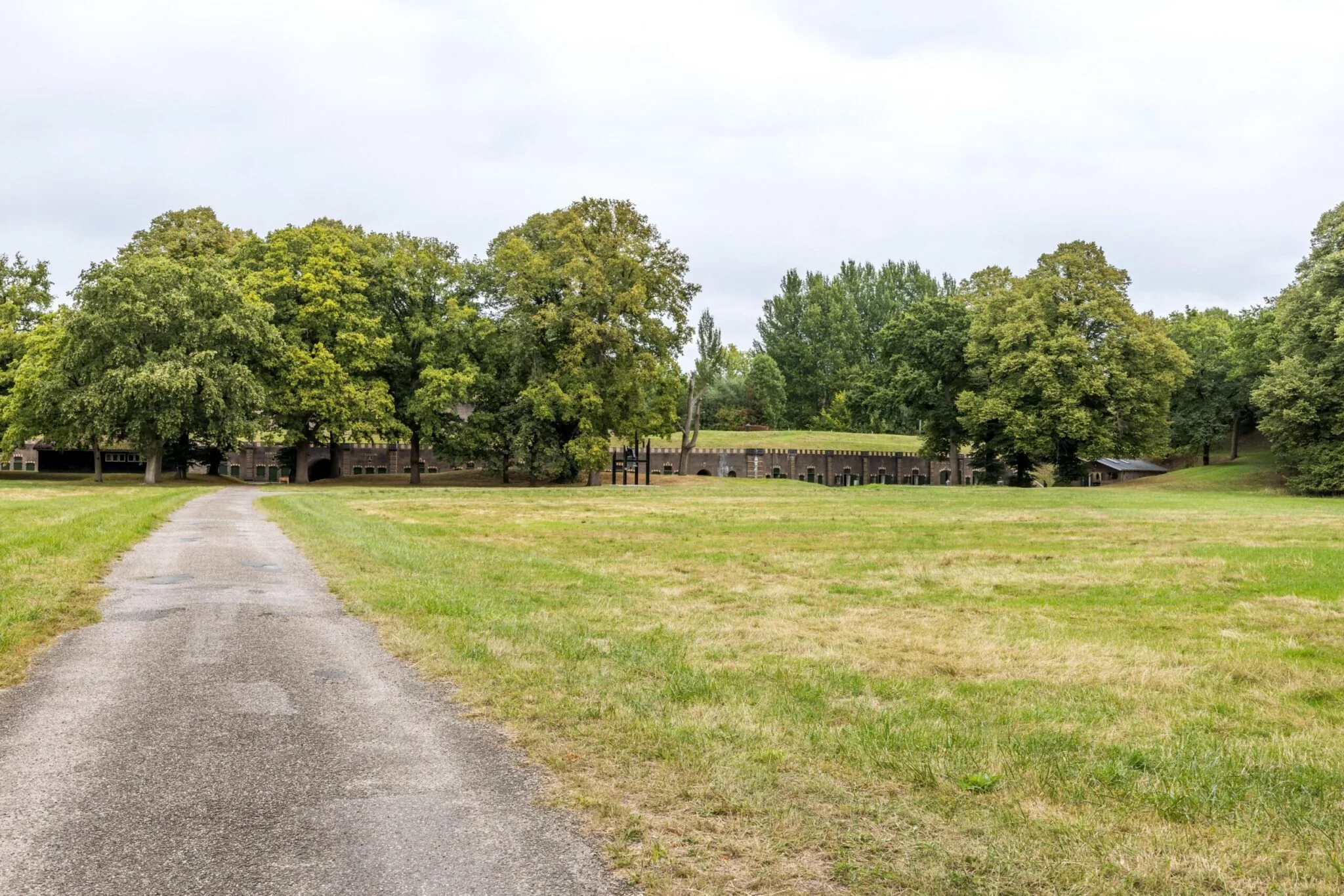 Asfaltweg leidt door een grasveld naar Fort bij Rijnauwen aan de Vossegatsedijk, omgeven door groene bomen.