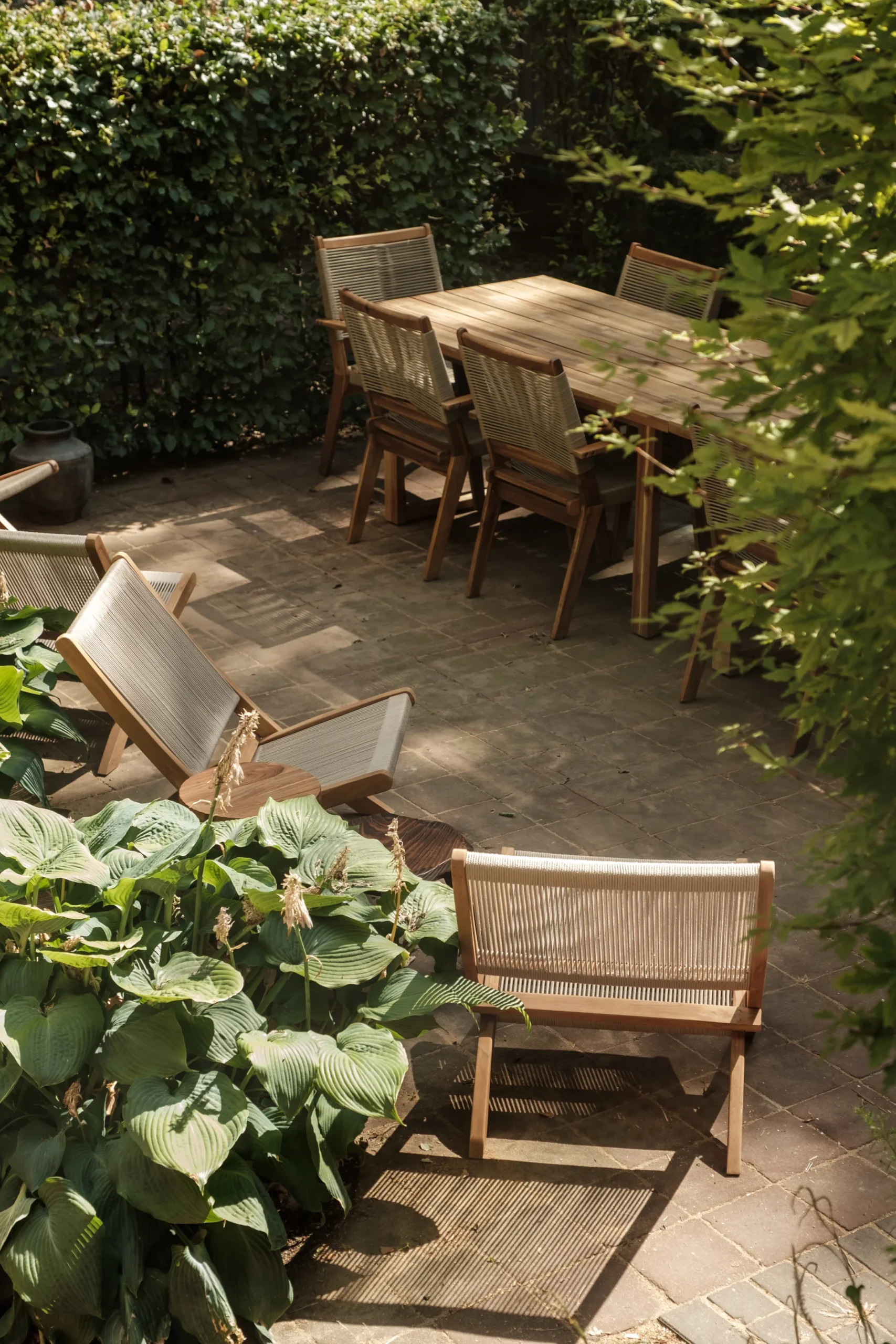 Tuin met houten tafel en zes stoelen omringd door groene planten.