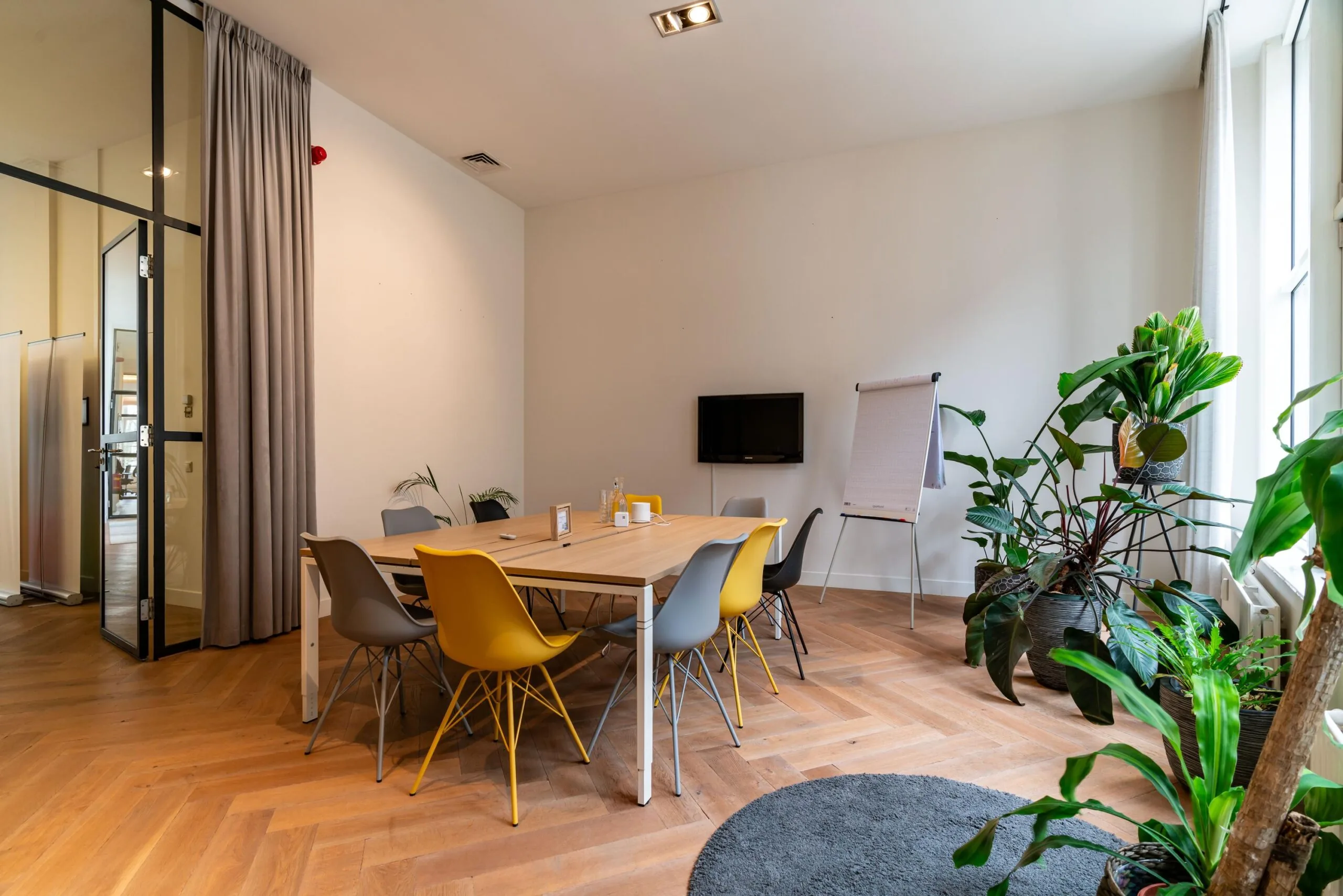 Vergaderruimte Lange Vijverberg met houten vloer, kleurrijke stoelen, flip-over en veel groene planten bij het raam.