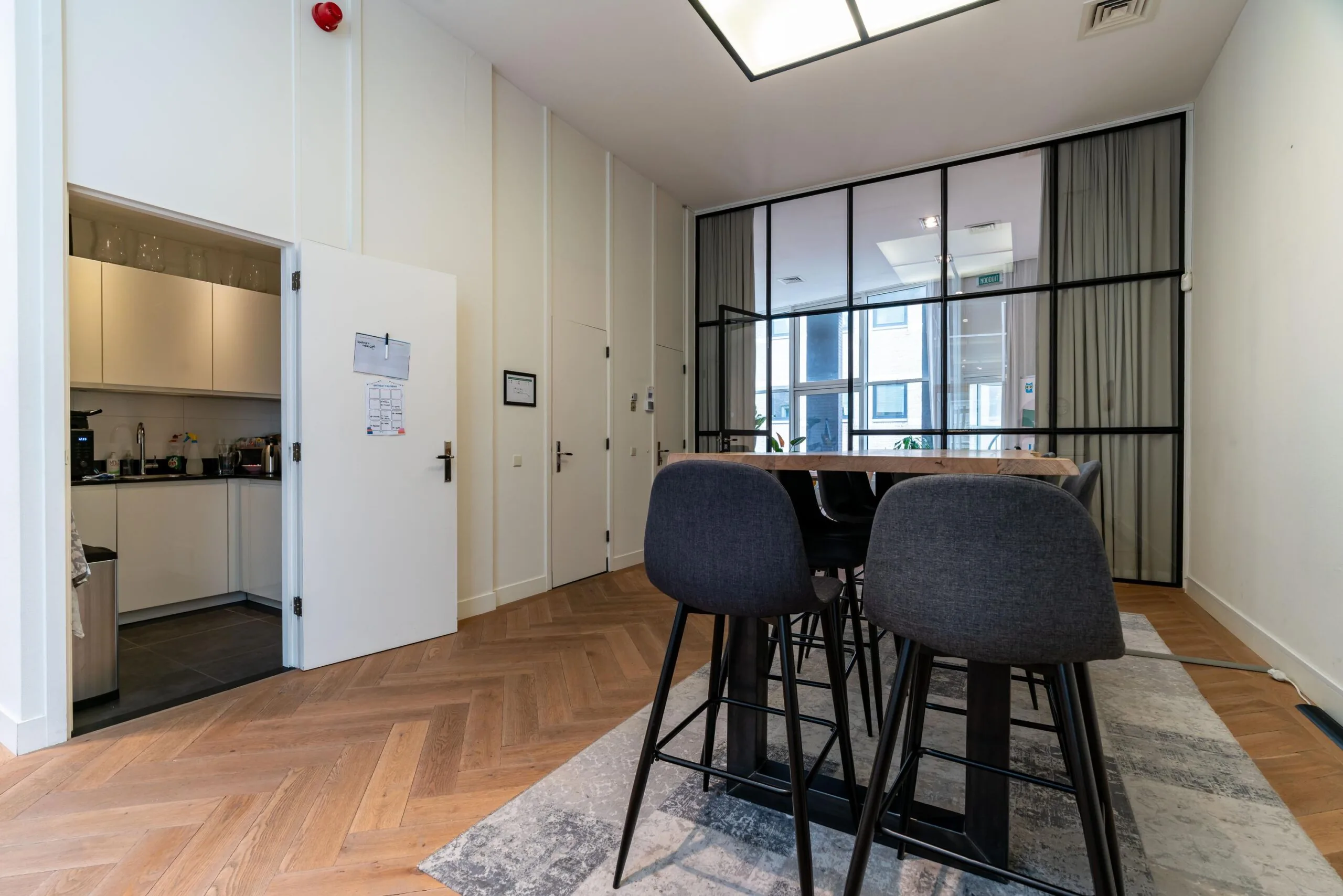 Vergaderruimte aan de Lange Vijverberg met hoge tafel, barkrukken en glazen scheidingswand naast een open deur naar de keuken.