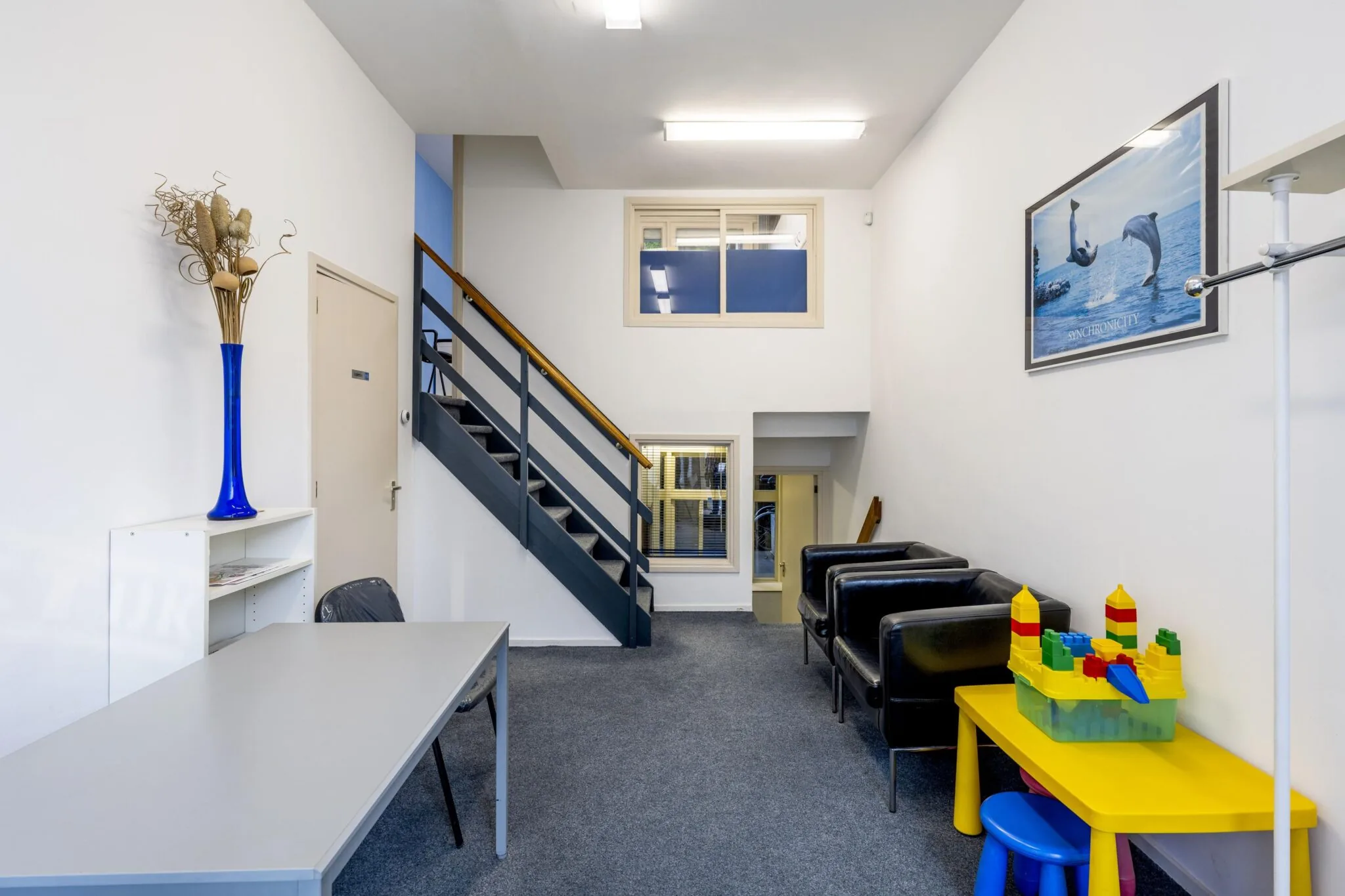 Wachtruimte met zwarte stoelen, een kinderspeelhoek, een trap en een bureau in een kantoor aan de Overtoom.