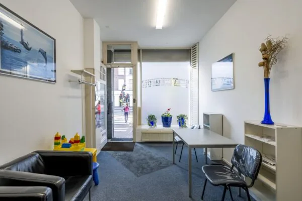 Wachtruimte met eenvoudige inrichting, kinderspeelhoek en uitzicht op de straat bij Overtoom 175.