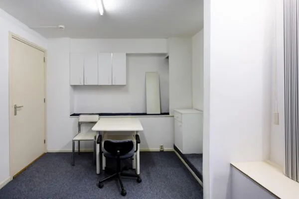 Kleine, eenvoudig ingerichte werk- of vergaderruimte met twee stoelen, een tafel, een spiegel en witte kasten aan de muur.