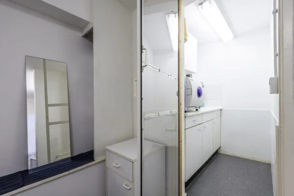 Kleine witte behandel- of sterilisatieruimte met een kast, spiegel en medisch apparaat op het aanrecht.