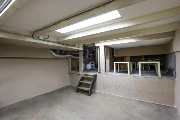 Kelderruimte met betonnen vloer, lage plafonds, een trapje naar een verhoogd gedeelte en zicht op opgeslagen tafels en technische installaties.