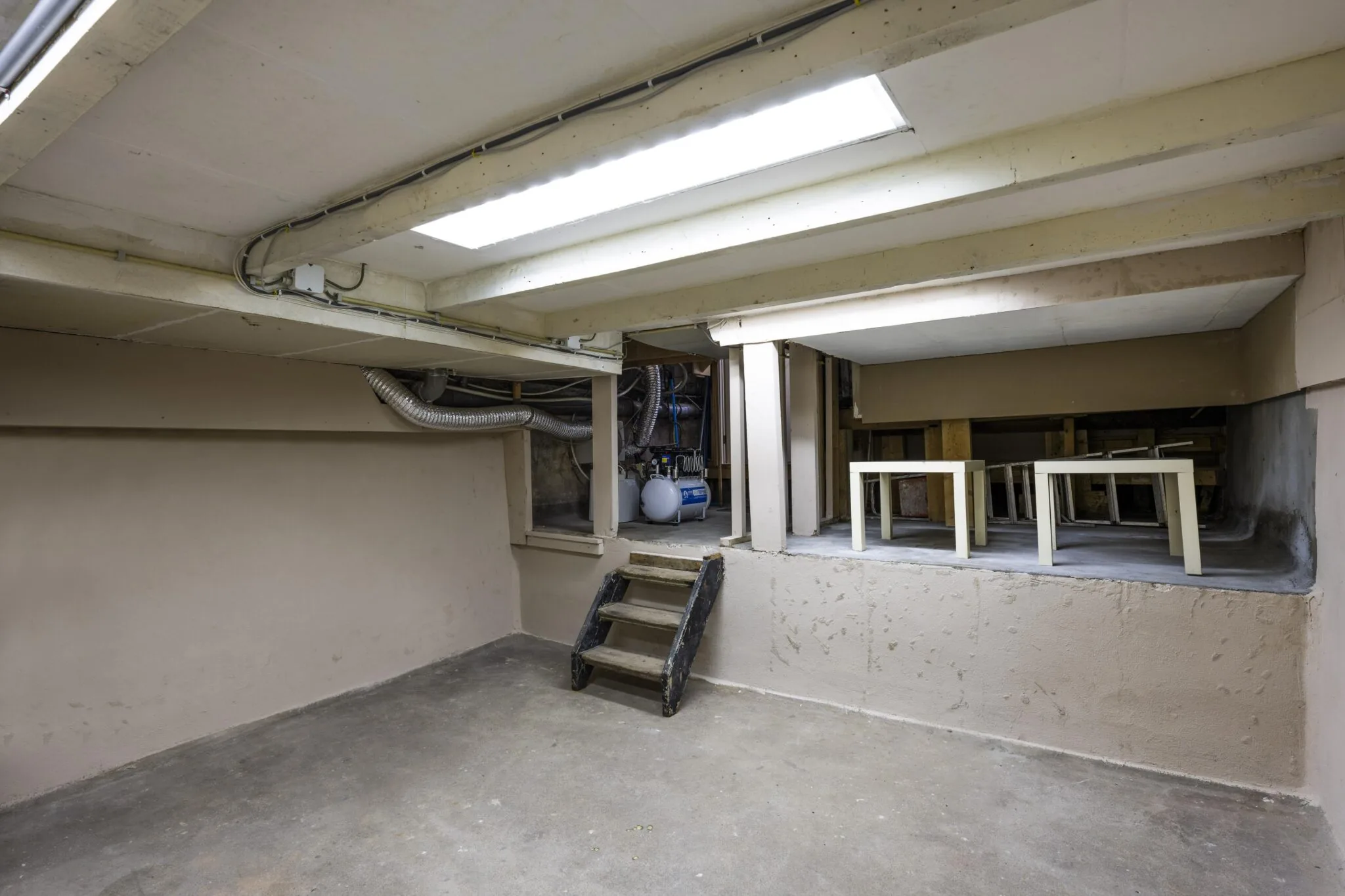 Kelderruimte met betonnen vloer, lage plafonds, een trapje naar een verhoogd gedeelte en zicht op opgeslagen tafels en technische installaties.