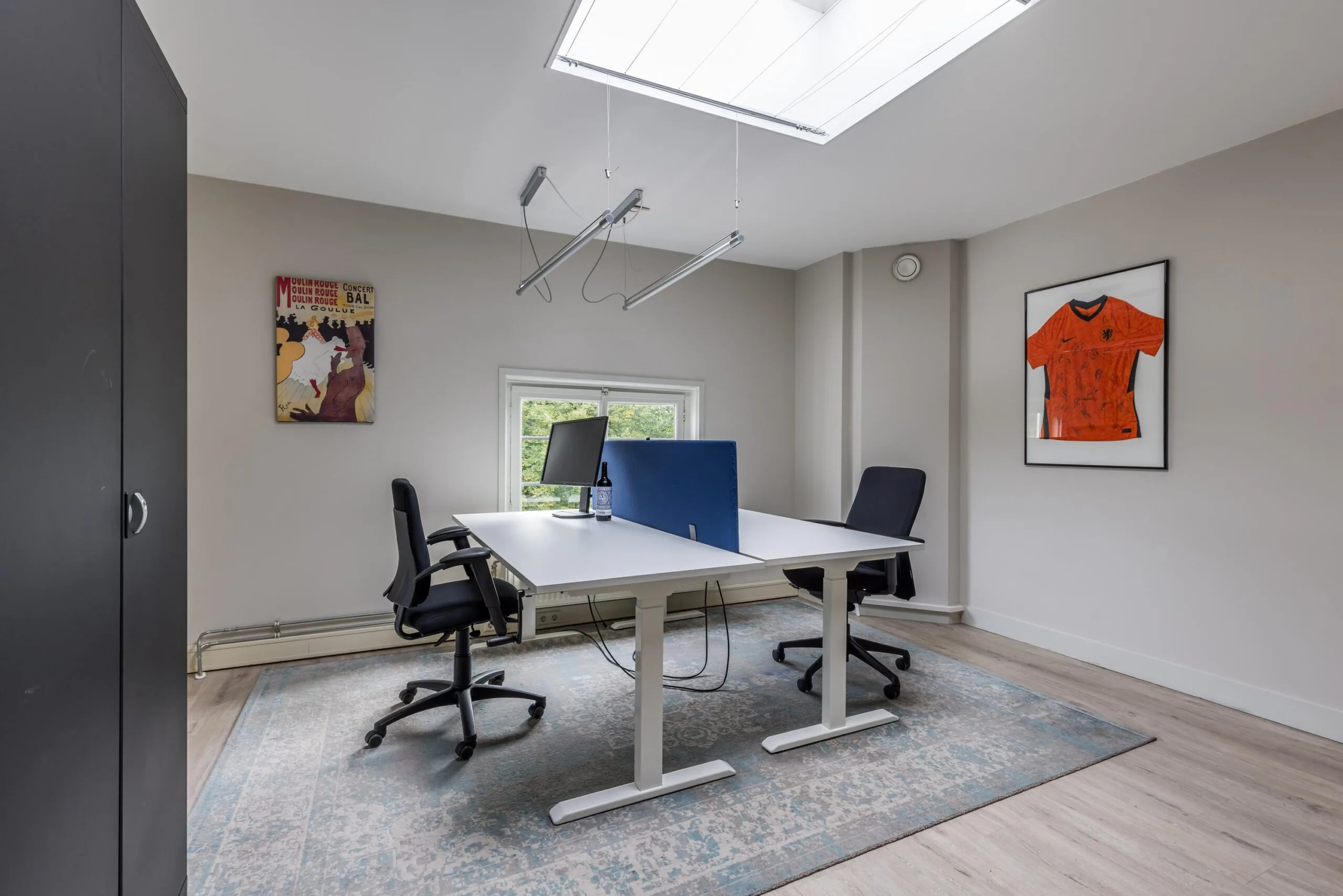 Modern ingericht kantoor met twee bureaustoelen, een gedeeld bureau, een computer en decoraties aan de muur, waaronder een ingelijst Nederlands voetbalshirt.