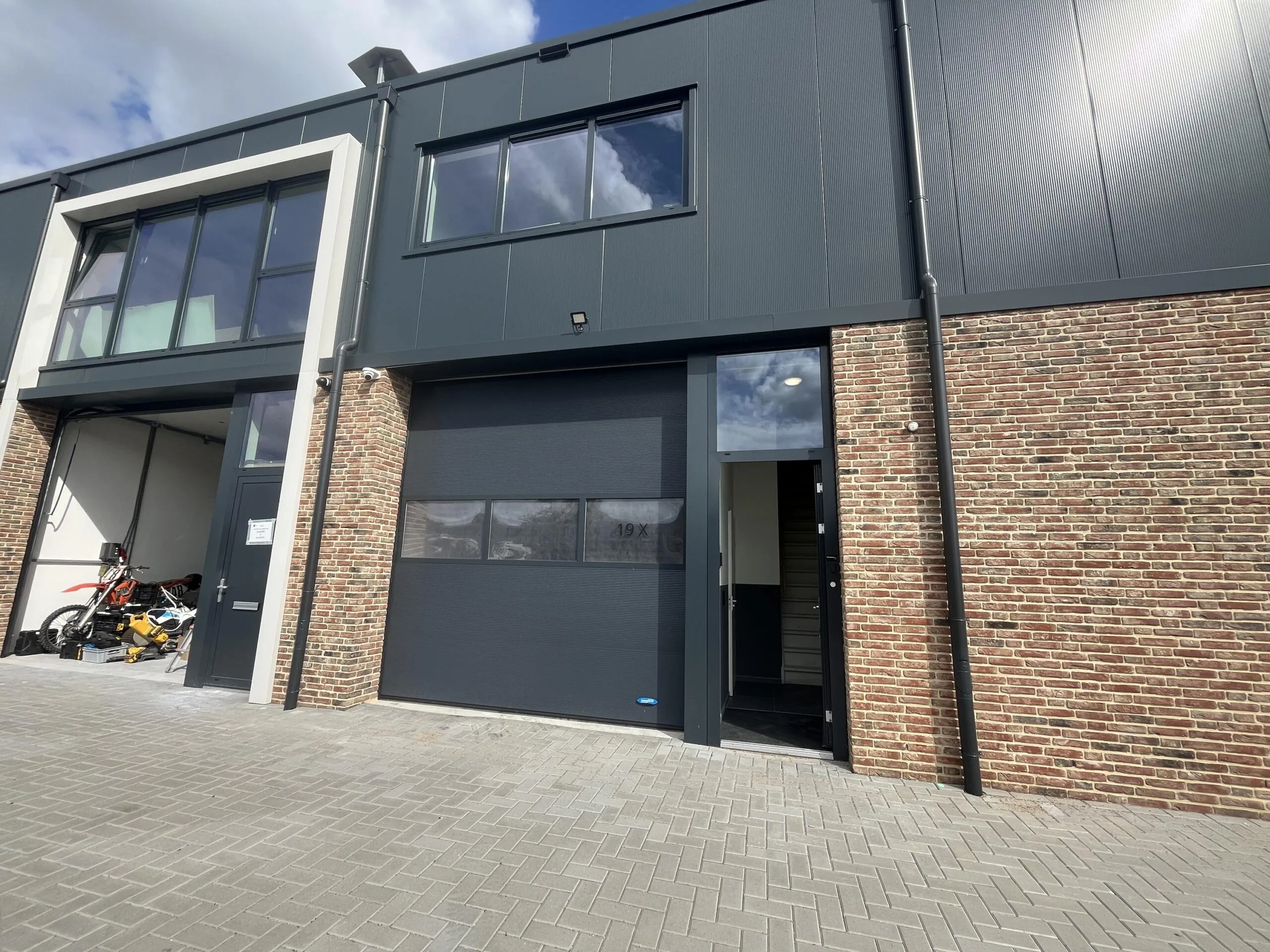 Een modern bedrijfsgebouw met een bakstenen gevel en een gesloten garagedeur, naast een open ruimte met een motorfiets erin.