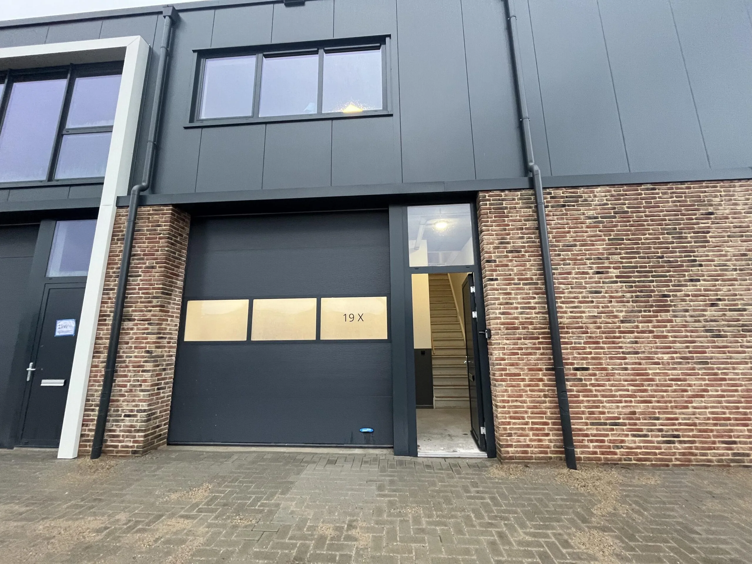 Een modern industrieel gebouw met een grote garagedeur en een openstaande deur naar een trap binnenin.