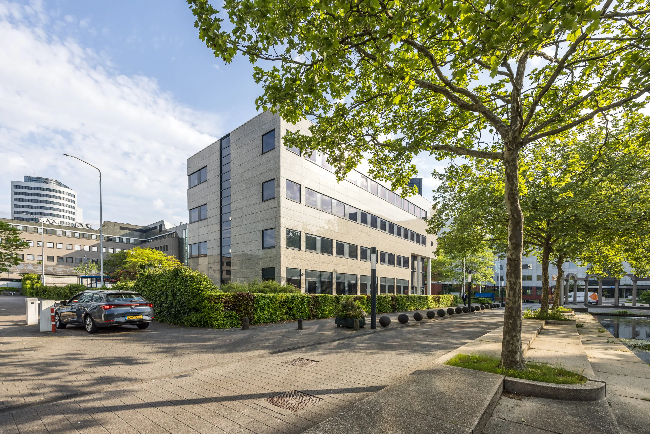 Kantoorgebouw aan de Varrolaan met bomen en bestrating op de voorgrond, op een zonnige dag.