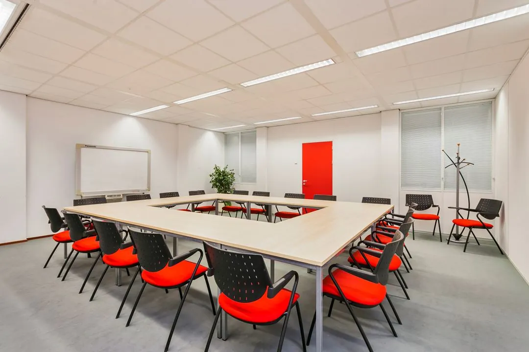 Vergaderruimte met U-vormige tafel, zwarte stoelen met rode zittingen, een whiteboard en een rode deur.