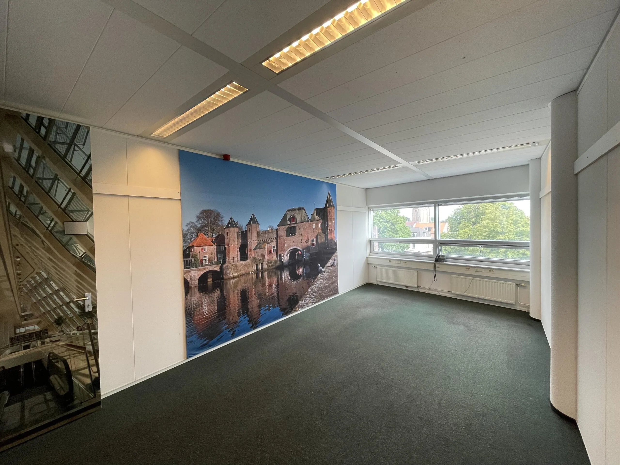 Leeg kantoor met groot muurfotobehang van de Koppelpoort in Amersfoort en uitzicht op bomen en gebouwen door de ramen.