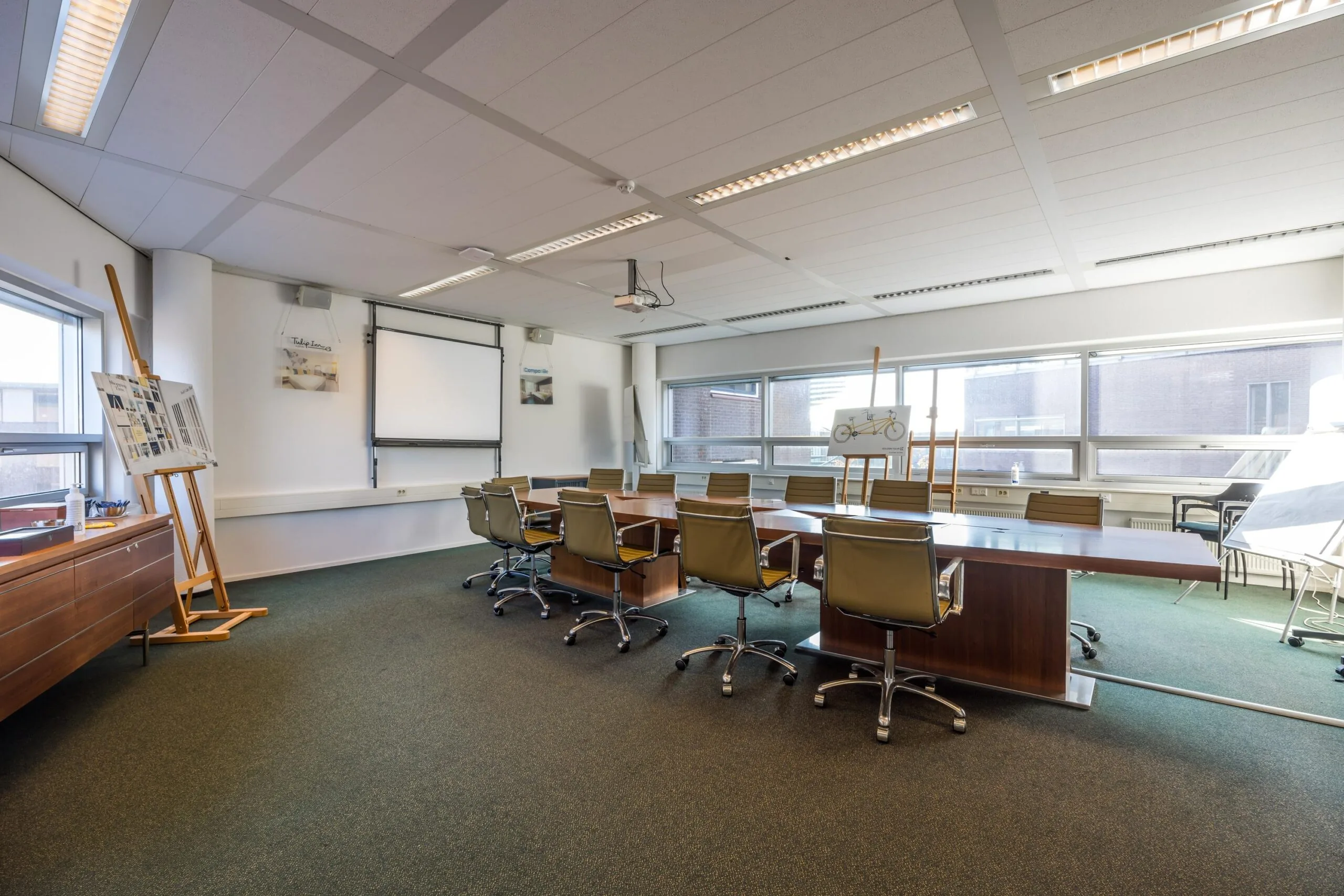 Vergaderruimte aan de Arnhemseweg met lange houten tafel, draaistoelen, whiteboard en tekenrekken bij de ramen.
