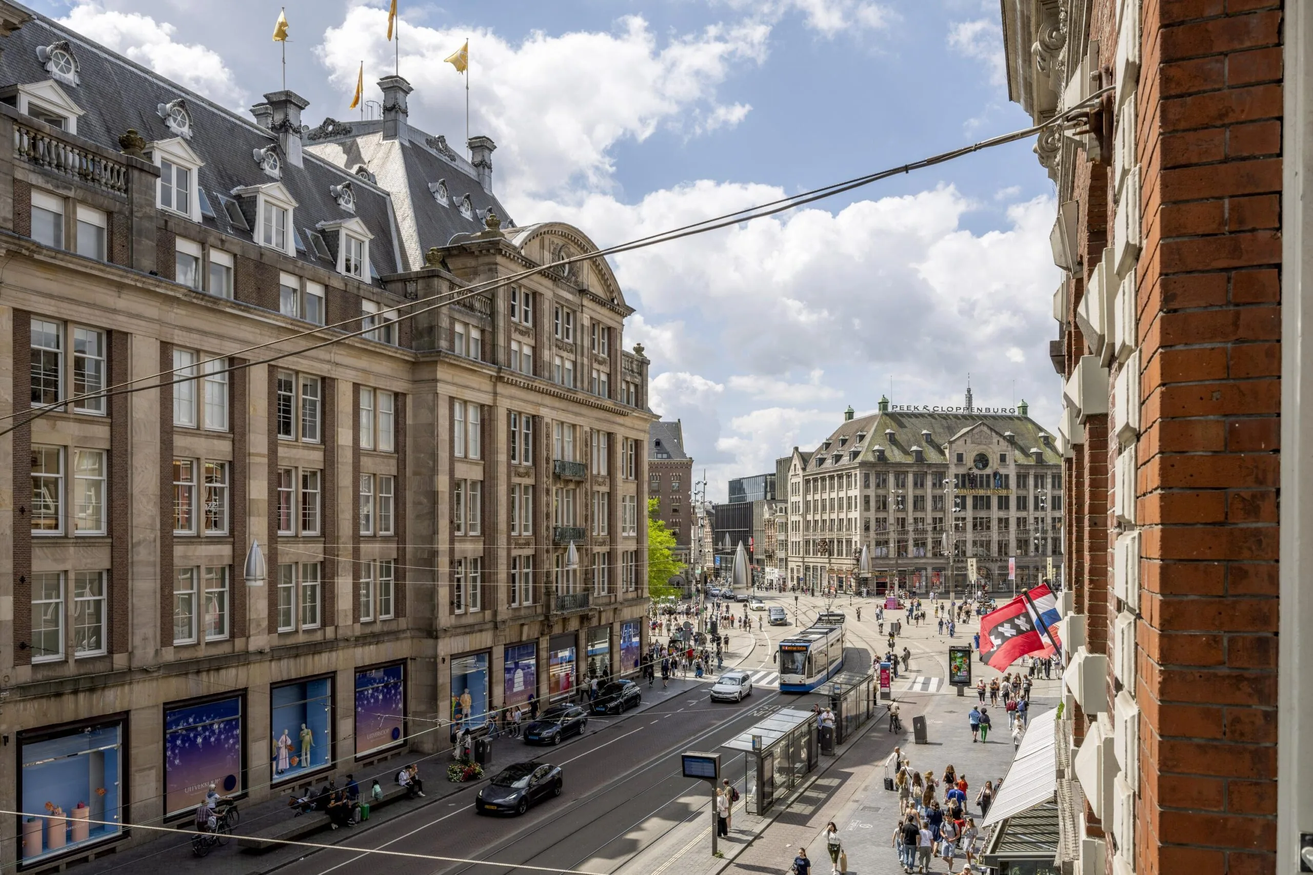 Drukke straat op het Damrak in Amsterdam met historische gebouwen, trams, winkelend publiek en het Peek & Cloppenburg-gebouw op de achtergrond.