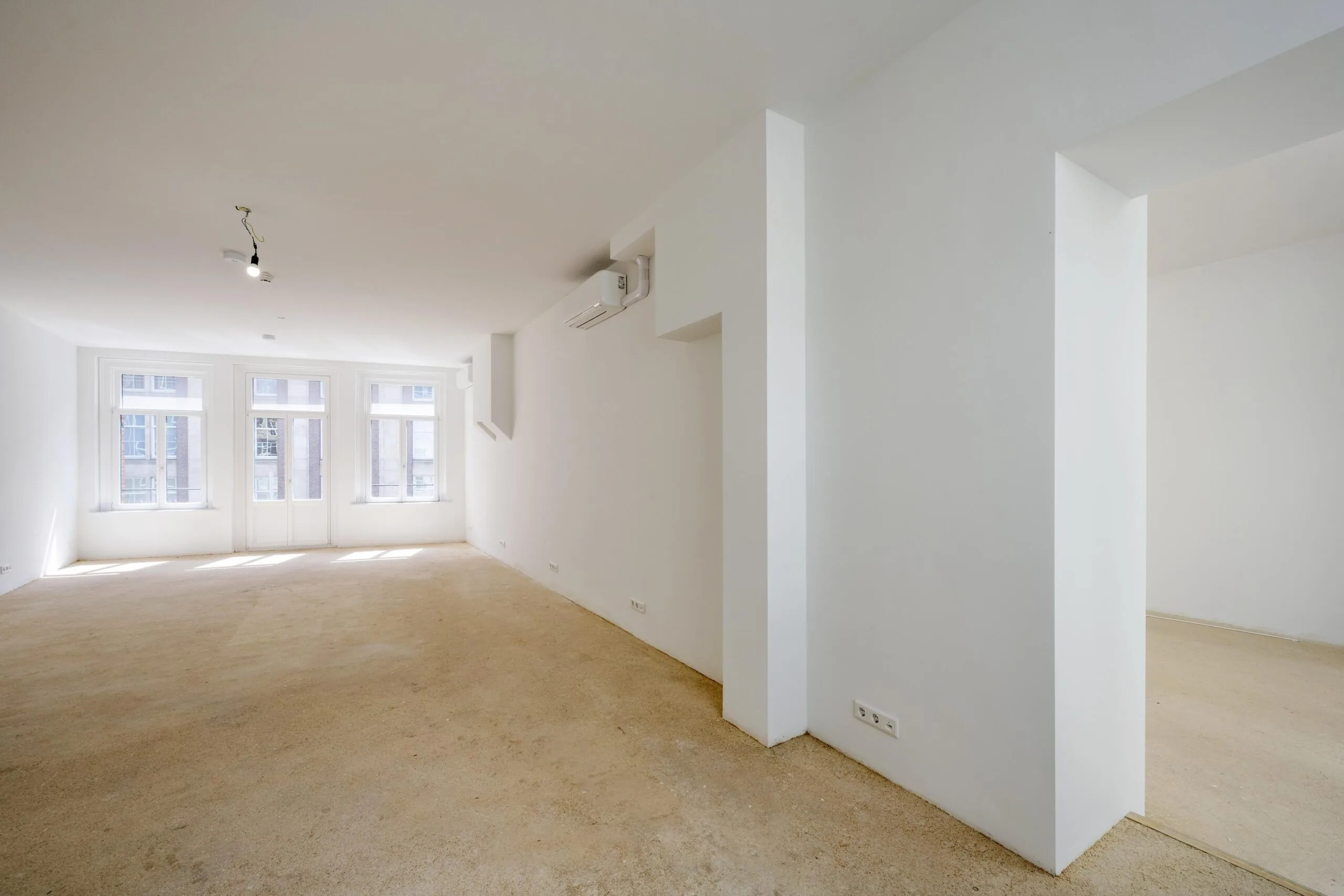 Leeg, gerenoveerd appartement aan het Damrak met witte muren, grote ramen en onafgewerkte vloer.