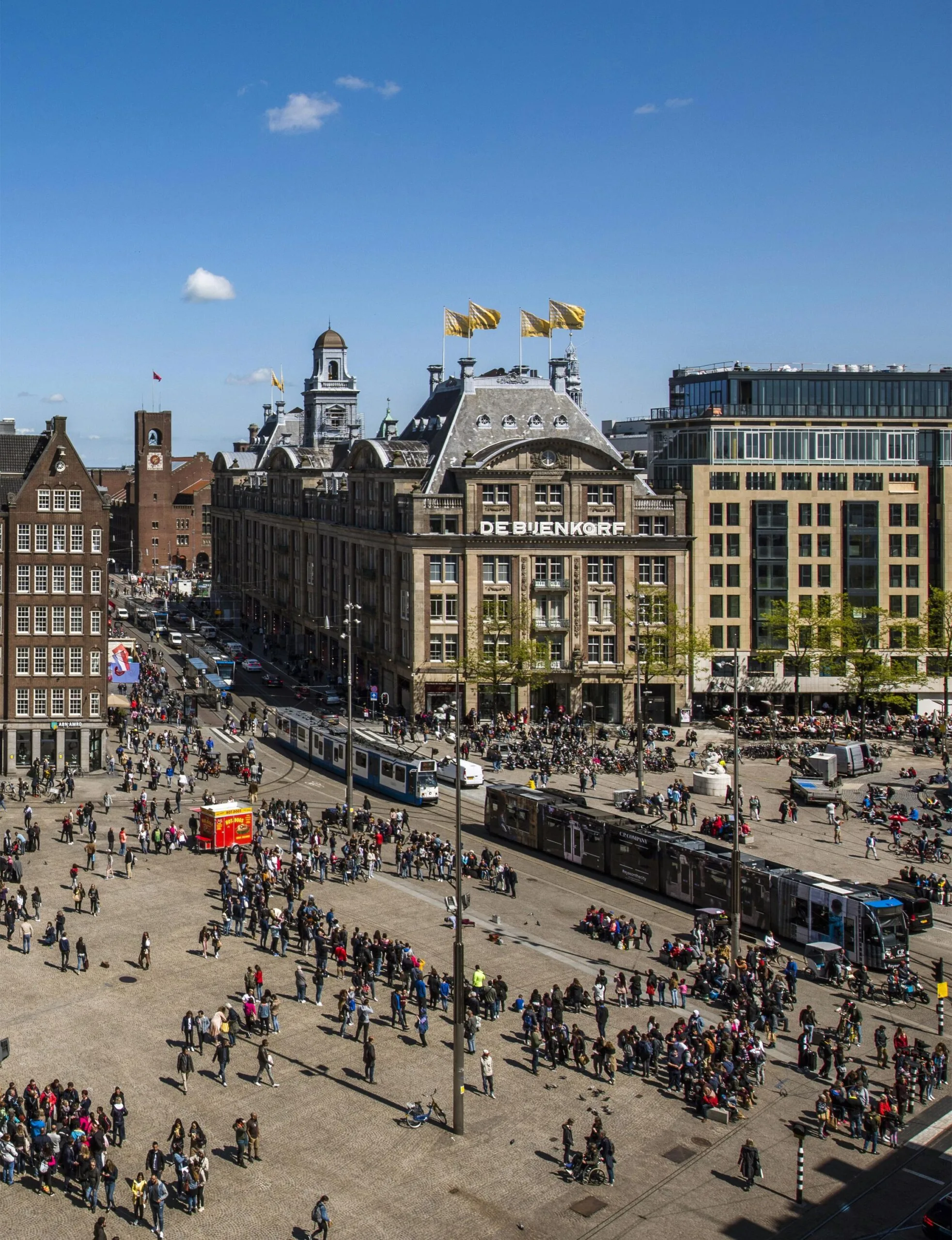 Drukke Damrak in Amsterdam met zicht op warenhuis De Bijenkorf en passerende trams op een zonnige dag.