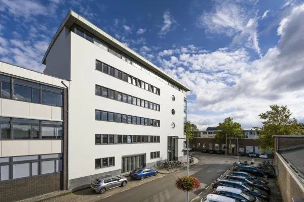 Modern office building on Helicopterstraat with parked cars and bicycles under a partly cloudy sky.