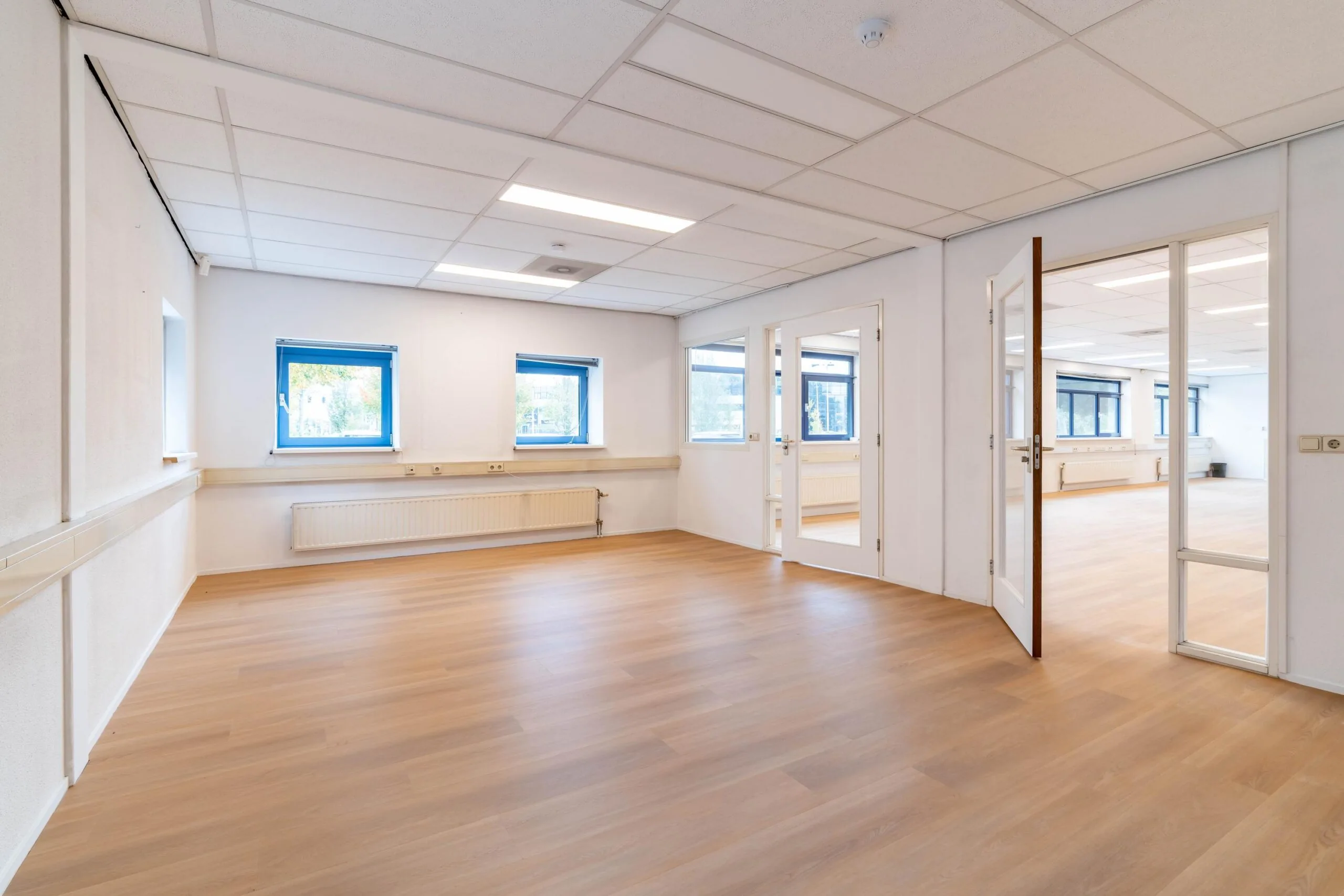 Lichte, lege kantoorruimte aan de Zuiderweg met houten vloer, systeemplafond en meerdere ramen en deuren.