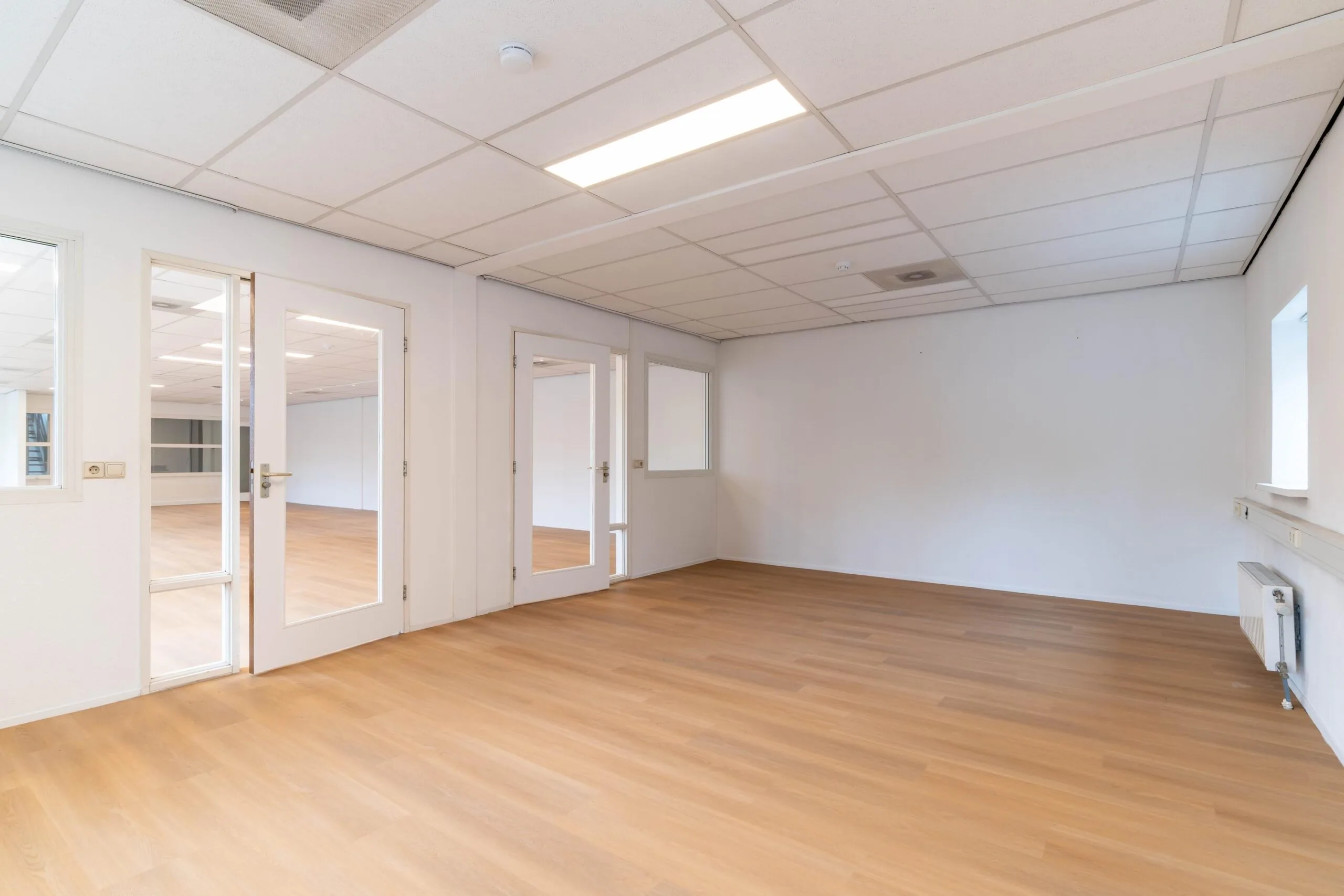 Lichte, lege kantoorruimte aan de Zuiderweg met houten vloer, systeemplafond en glazen binnendeuren.