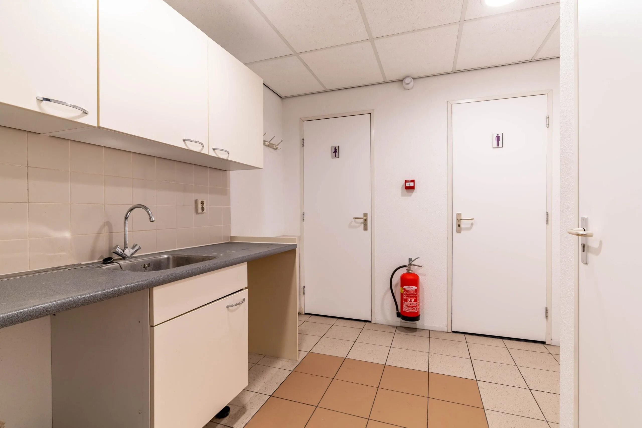 Kleine kitchenette met wasbak, twee toiletruimtes en een brandblusser aan de muur in een utilitaire ruimte op Zuiderweg.