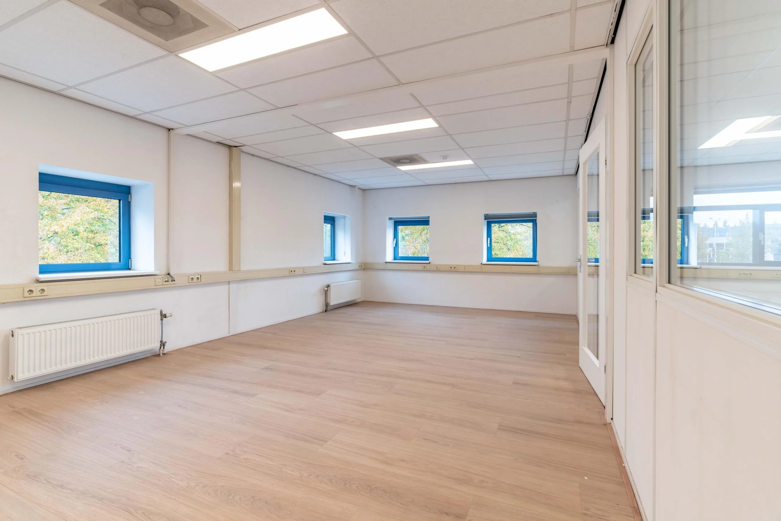 Lichte, lege kantoorruimte aan de Zuiderweg met houten vloer, systeemplafond en blauwe raamkozijnen.