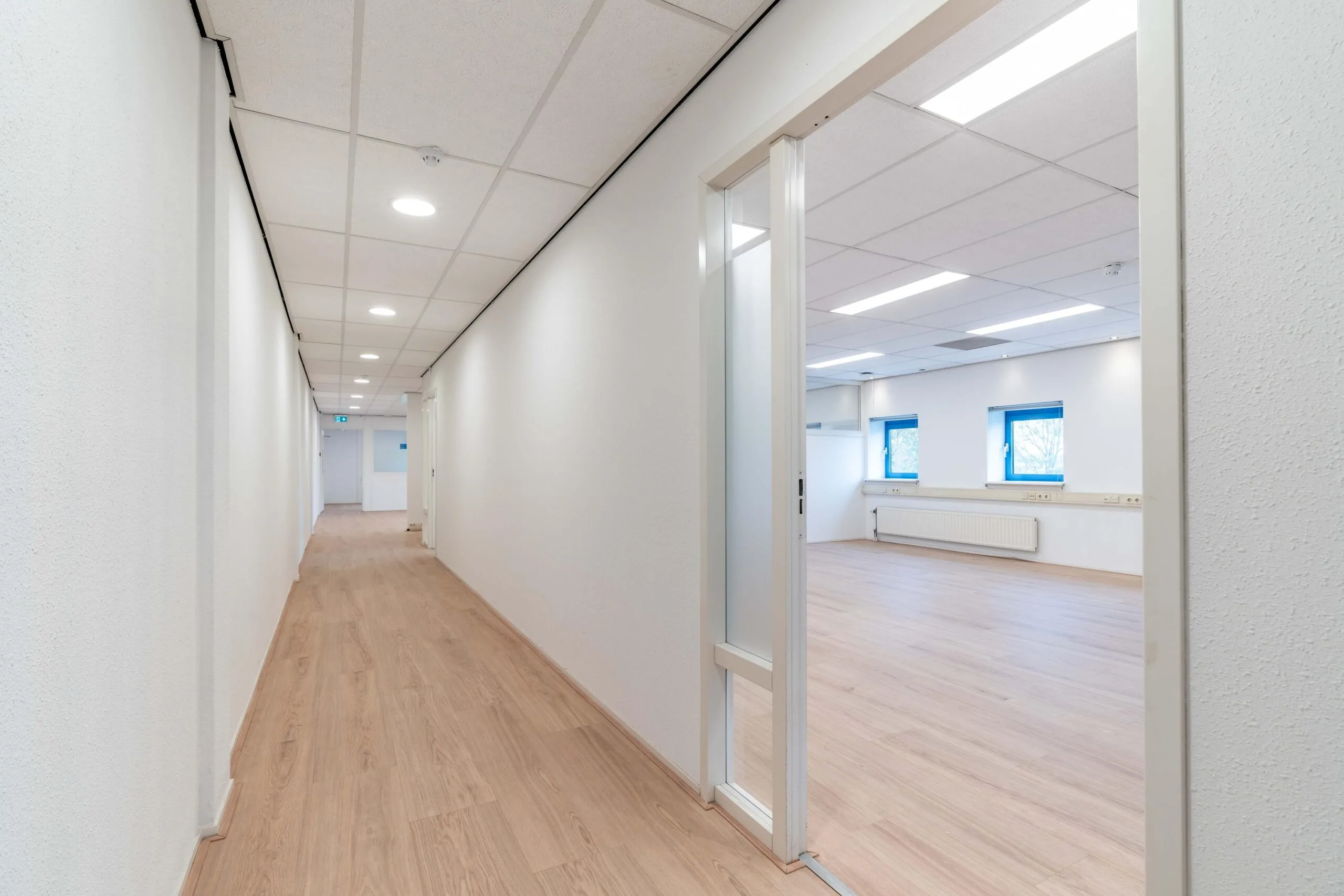 Lichte gang met houten vloer en een open deur naar een lege kantoorruimte aan de Zuiderweg.