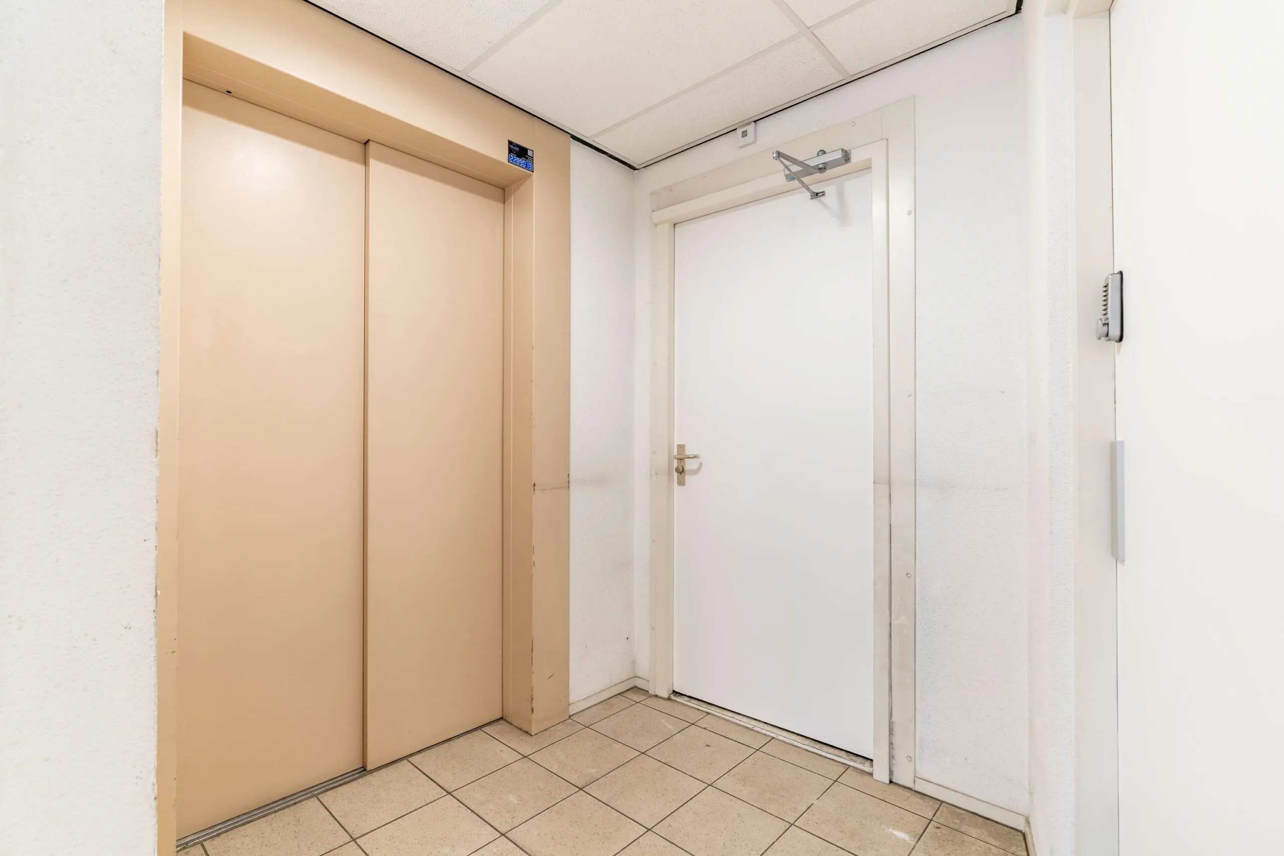 Binnenhal met een beige liftdeur en een witte deur met deurdranger in een pand aan de Zuiderweg.