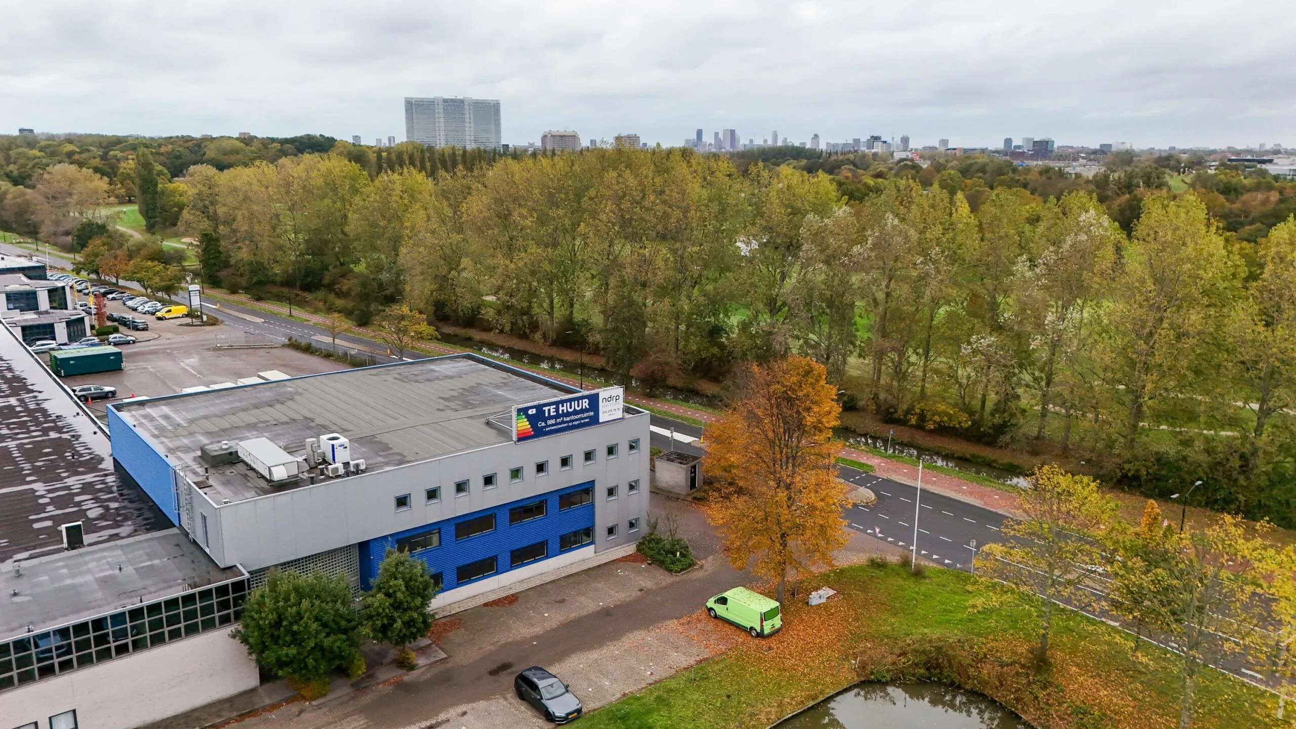 Bedrijfsgebouw aan de Zuiderweg met "Te Huur"-bord, gelegen naast een weg en omringd door bomen in herfstkleuren.