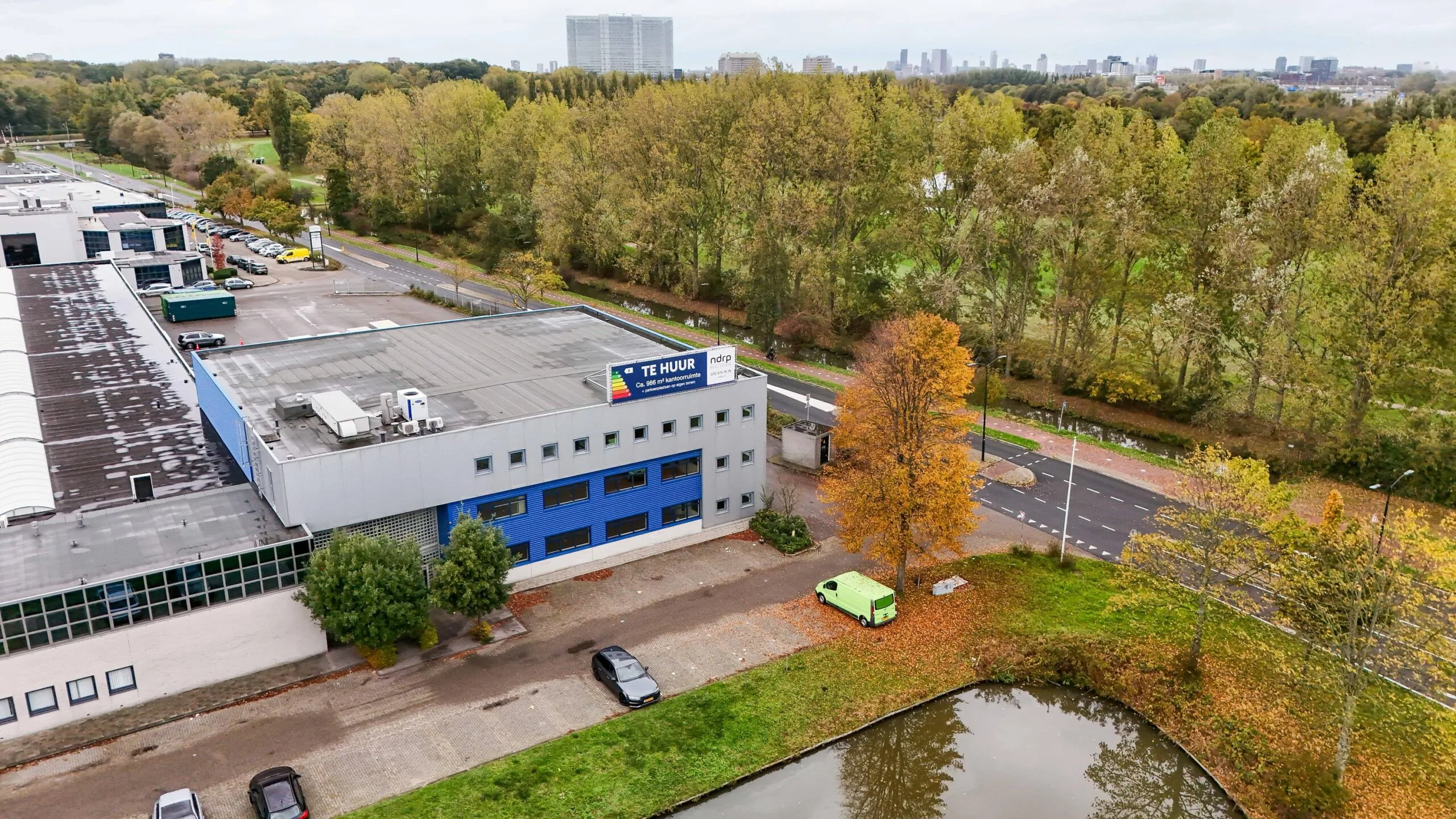 Luchtfoto van een kantoorpand aan de Zuiderweg met een "Te Huur"-bord, omringd door bomen en gelegen aan een vijver en een weg.
