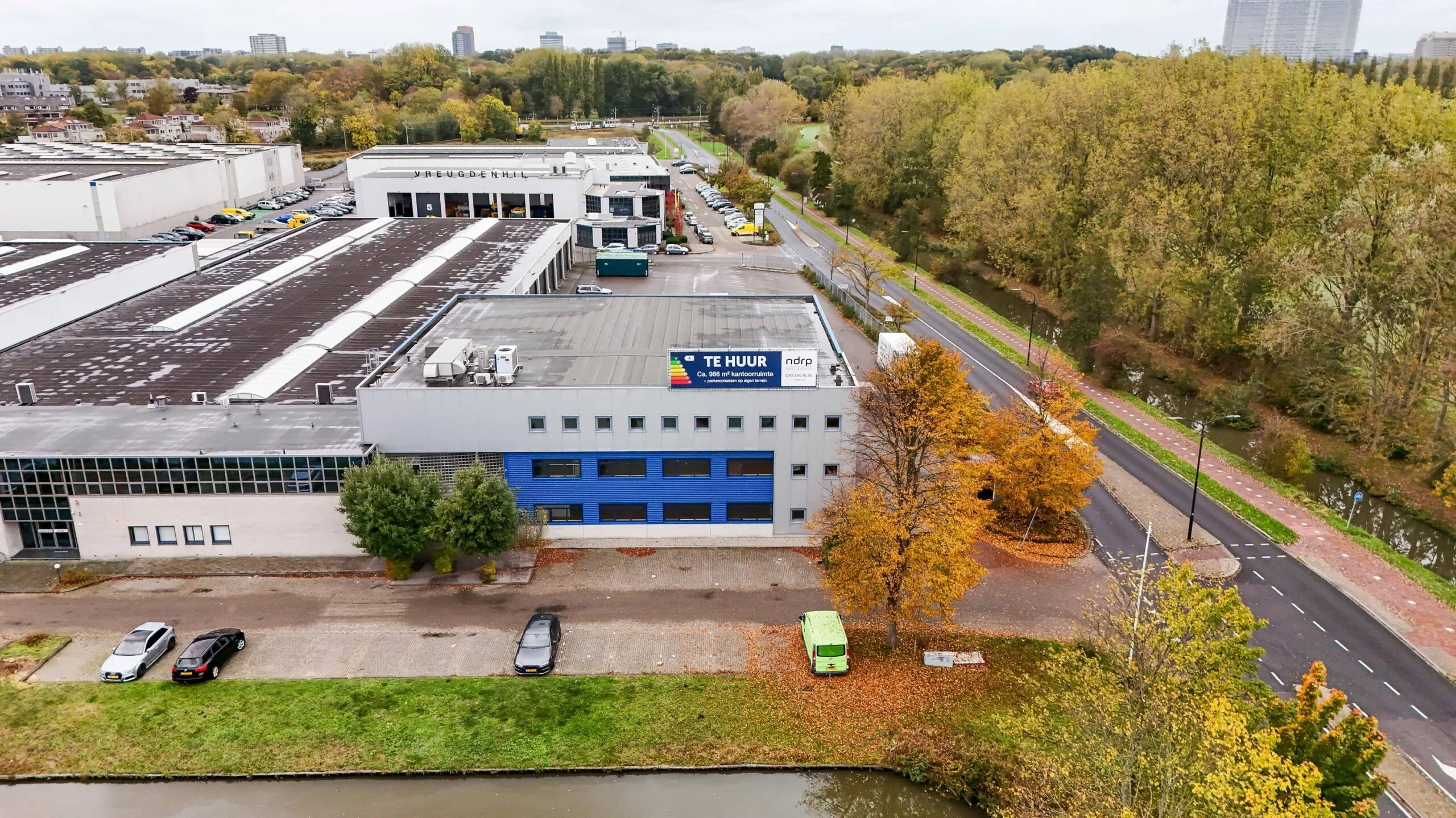 Luchtfoto van een bedrijfsgebouw aan de Zuiderweg met een "Te Huur"-bord op het dak, gelegen naast een weg en waterpartij.