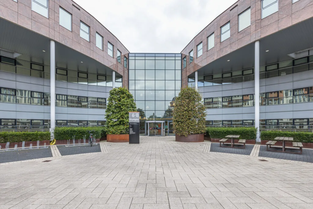 Modern office building entrance at Olympia 6-8 with large glass façade and surrounding landscaping.