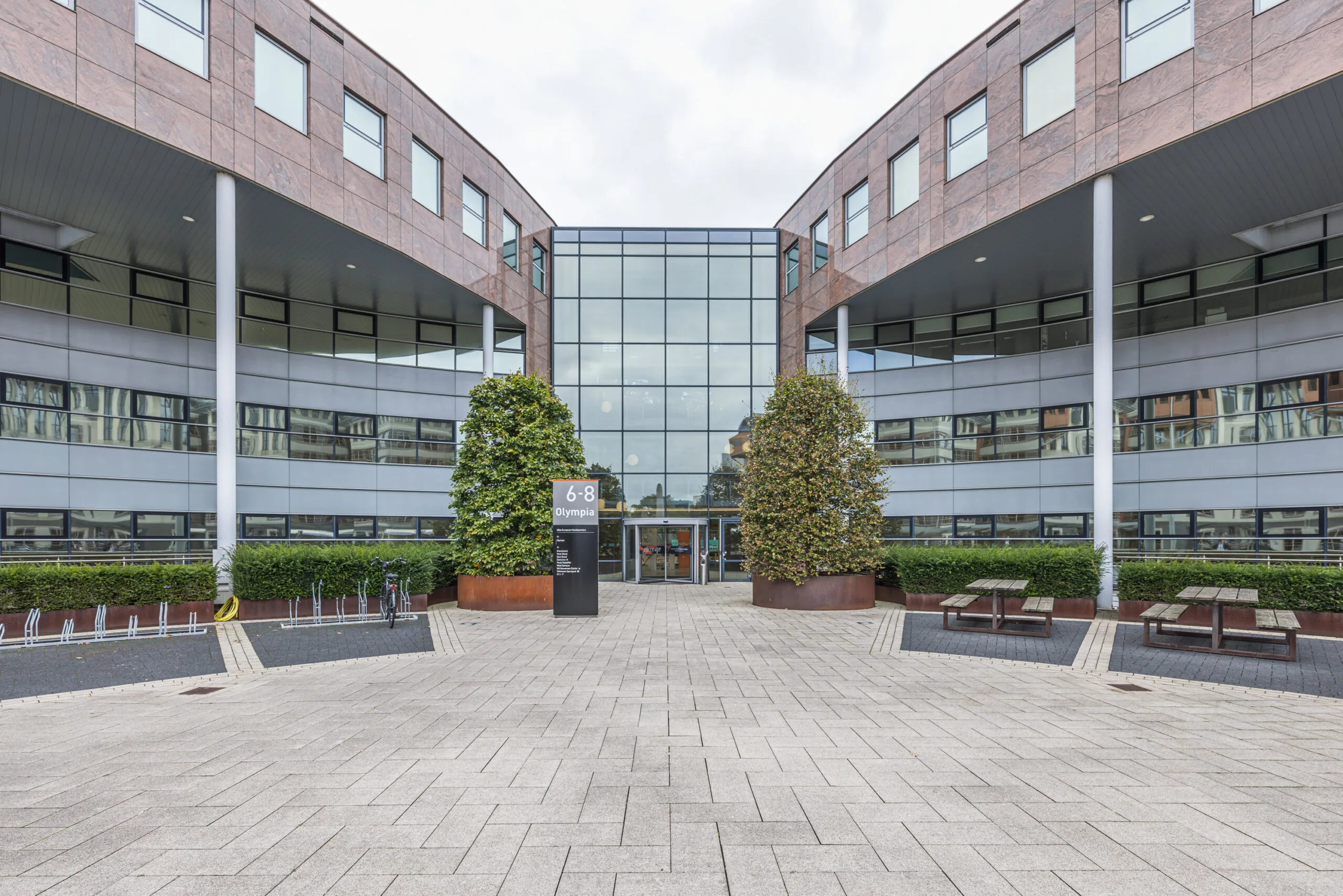 Modern office building entrance at Olympia 6-8 with large glass façade and surrounding landscaping.