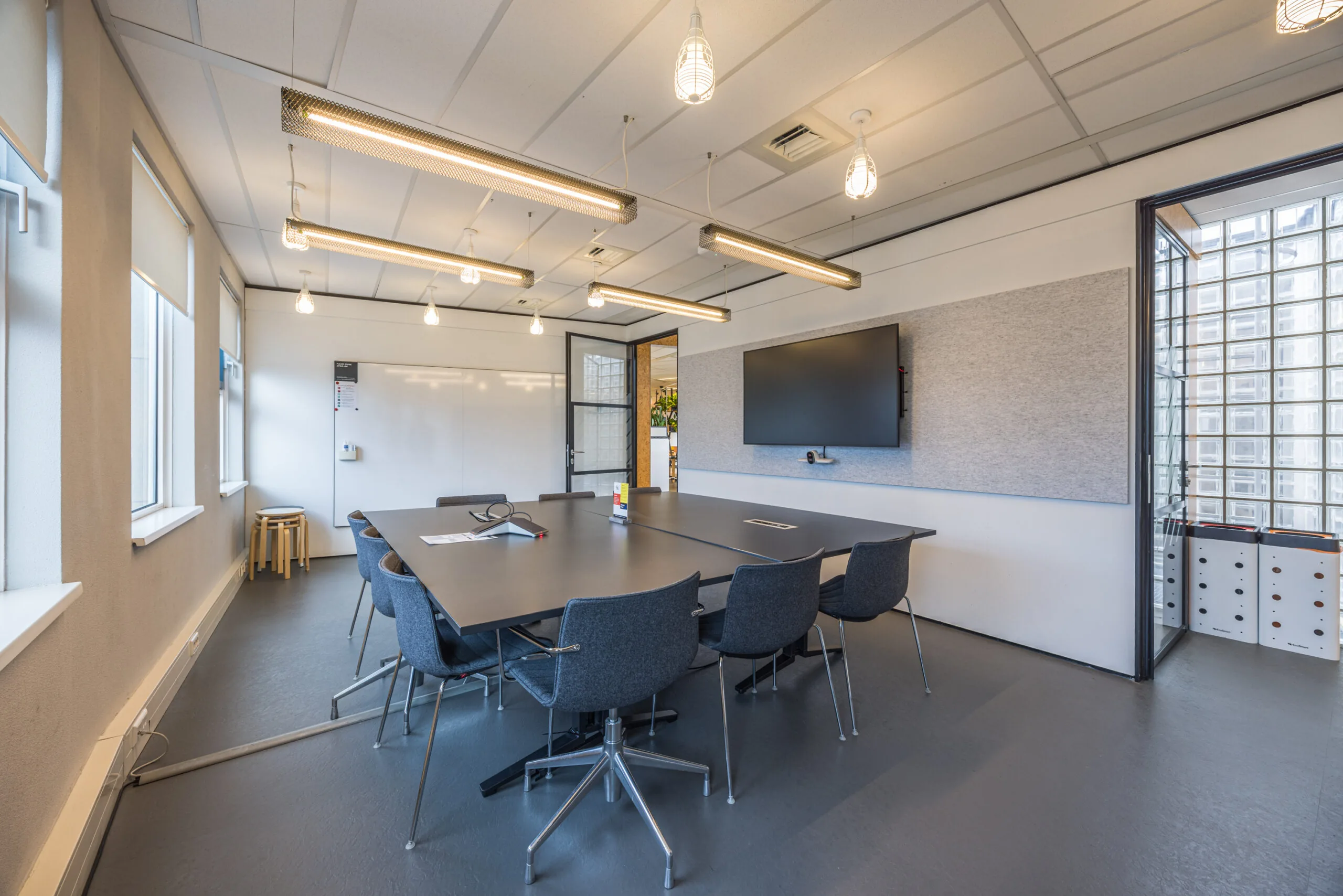 Modern conference room with a large meeting table, office chairs, a wall-mounted screen, and natural light from multiple windows.
