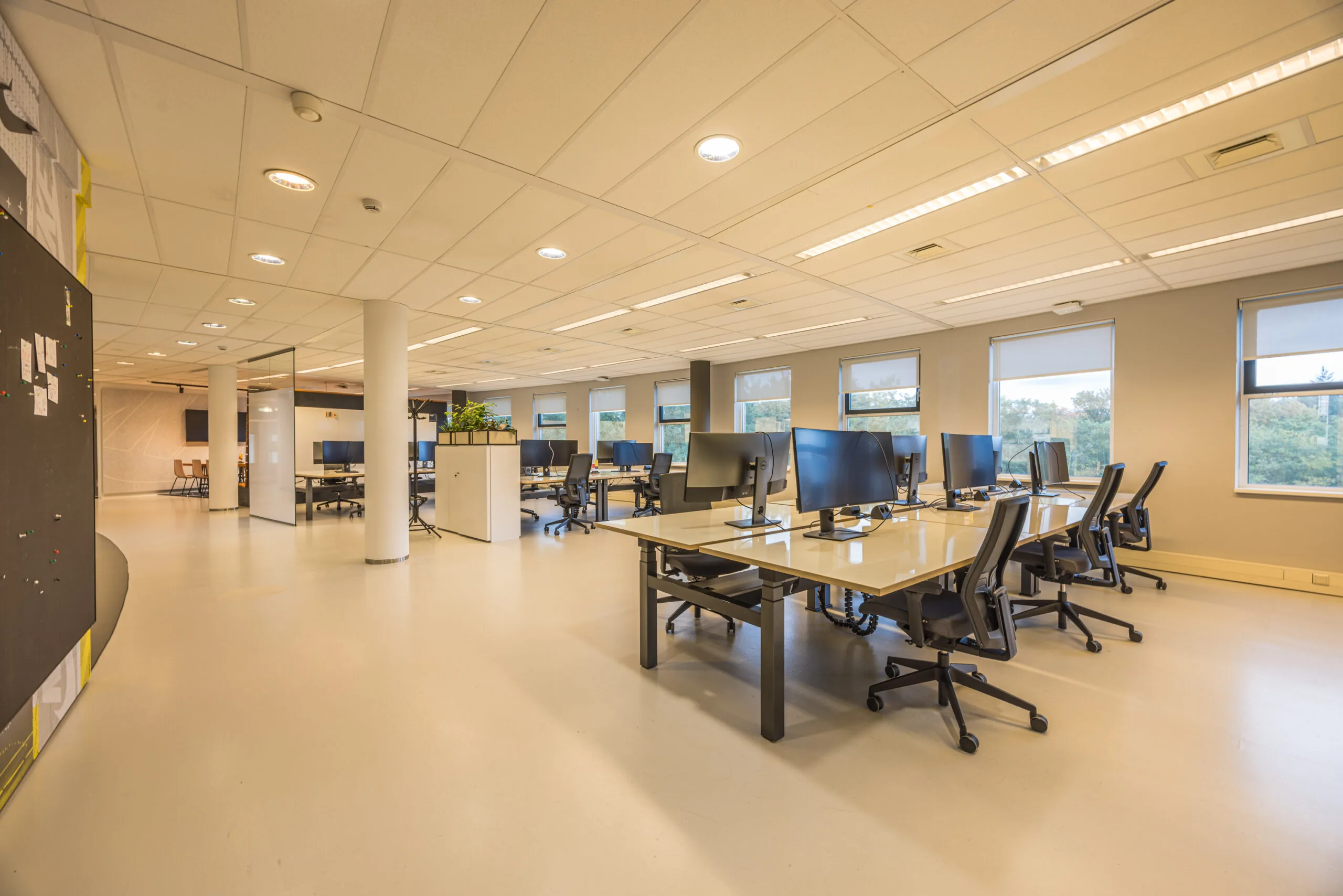 Modern open-plan office space with multiple desks, computer monitors, and large windows providing natural light.