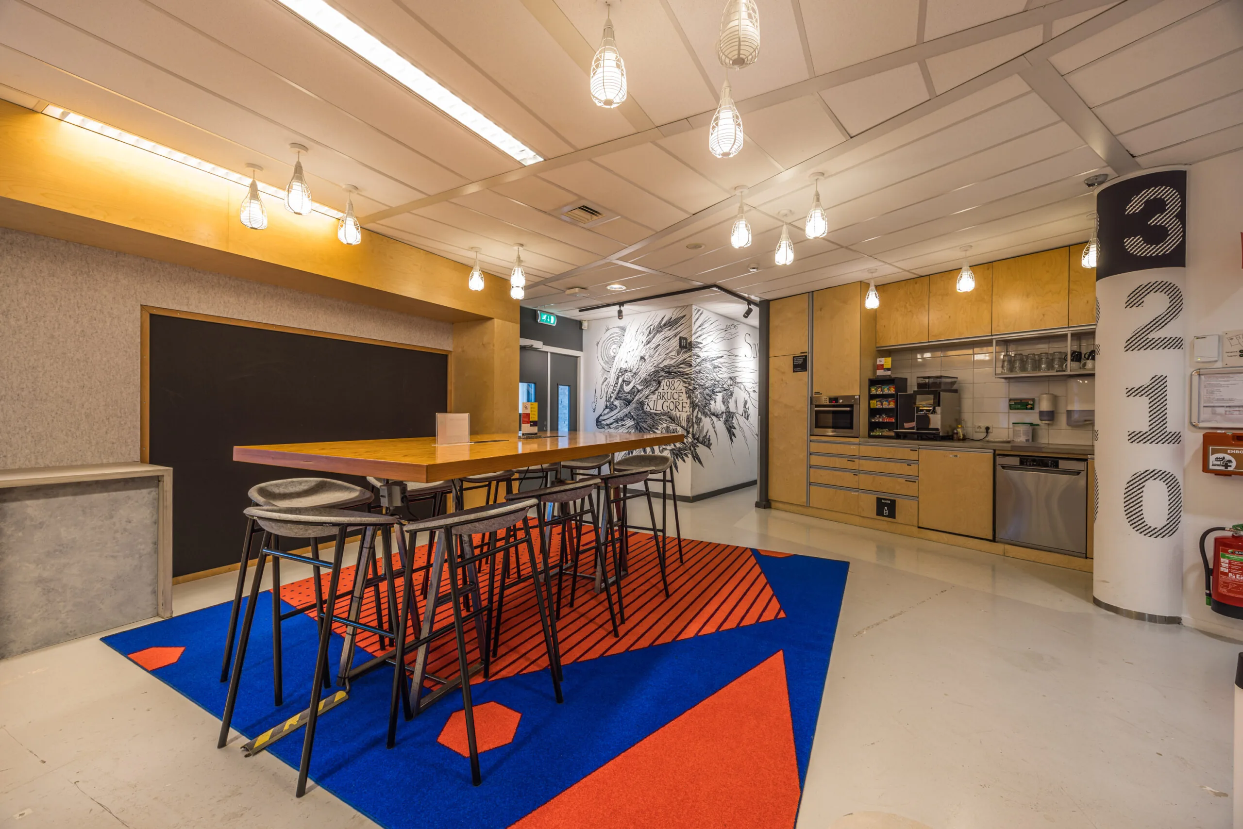 Modern office kitchen and break area with a long wooden table, colorful geometric carpet, wall art, and built-in appliances.